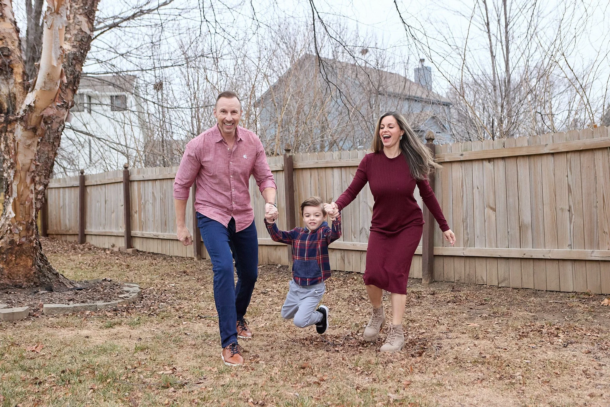 A family of three joyfully running and holding hands in a backyard with a wooden fence and leafless trees.
