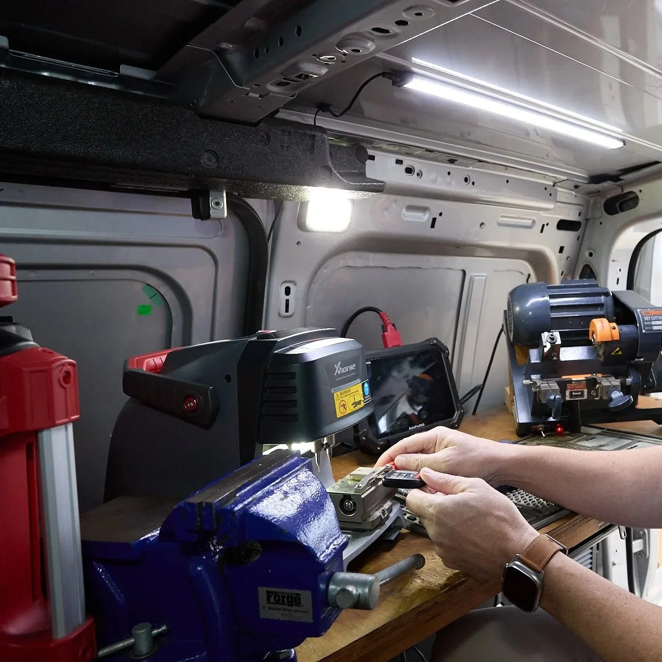 Inside a workshop or mobile workspace, a person with a wristwatch is working with a small device at a cluttered wooden workbench, surrounded by various tools and equipment, with shelving and a fluorescent light overhead.