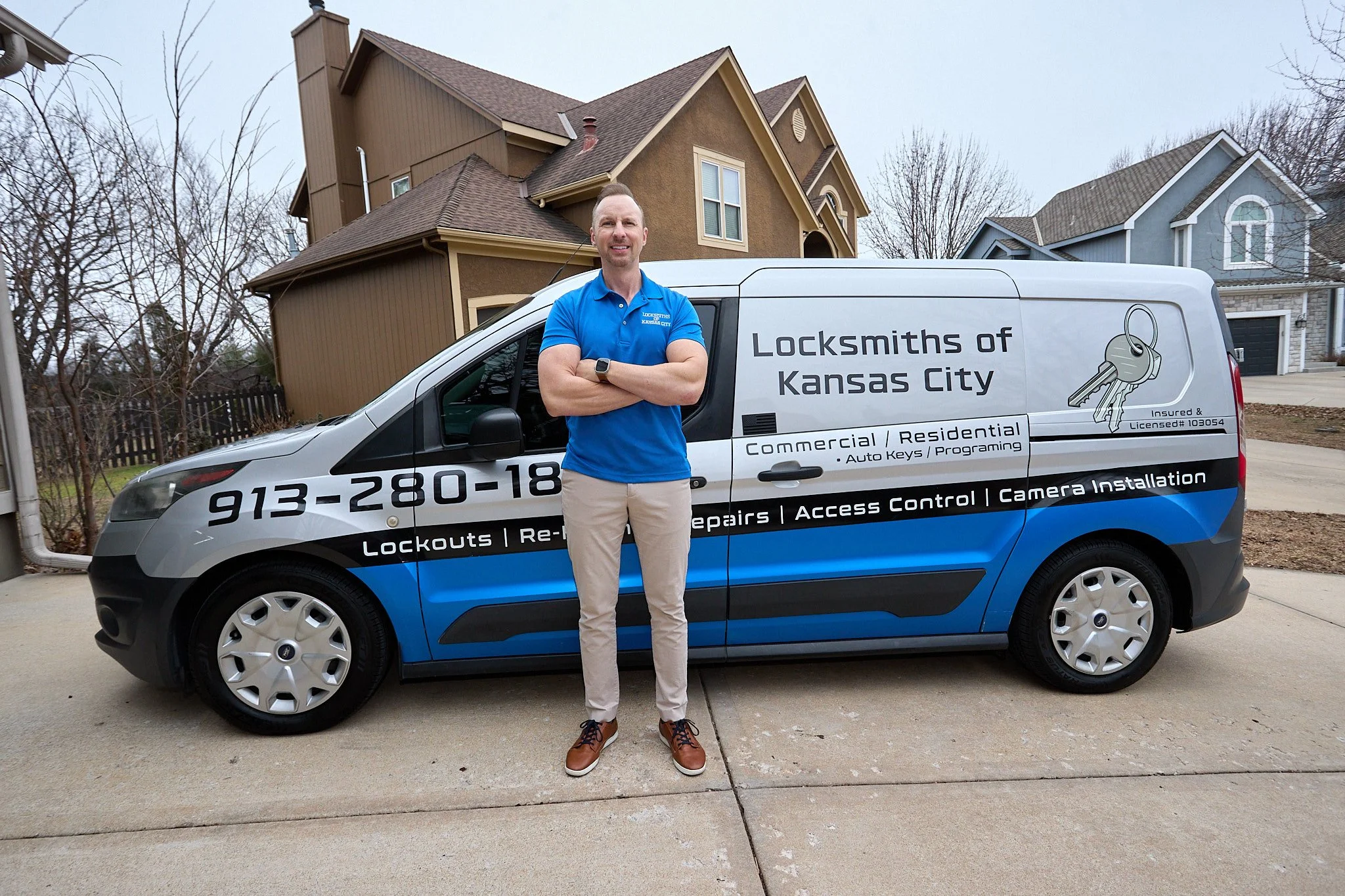 A man standing with arms crossed next to a branded locksmith service van in front of a suburban house.