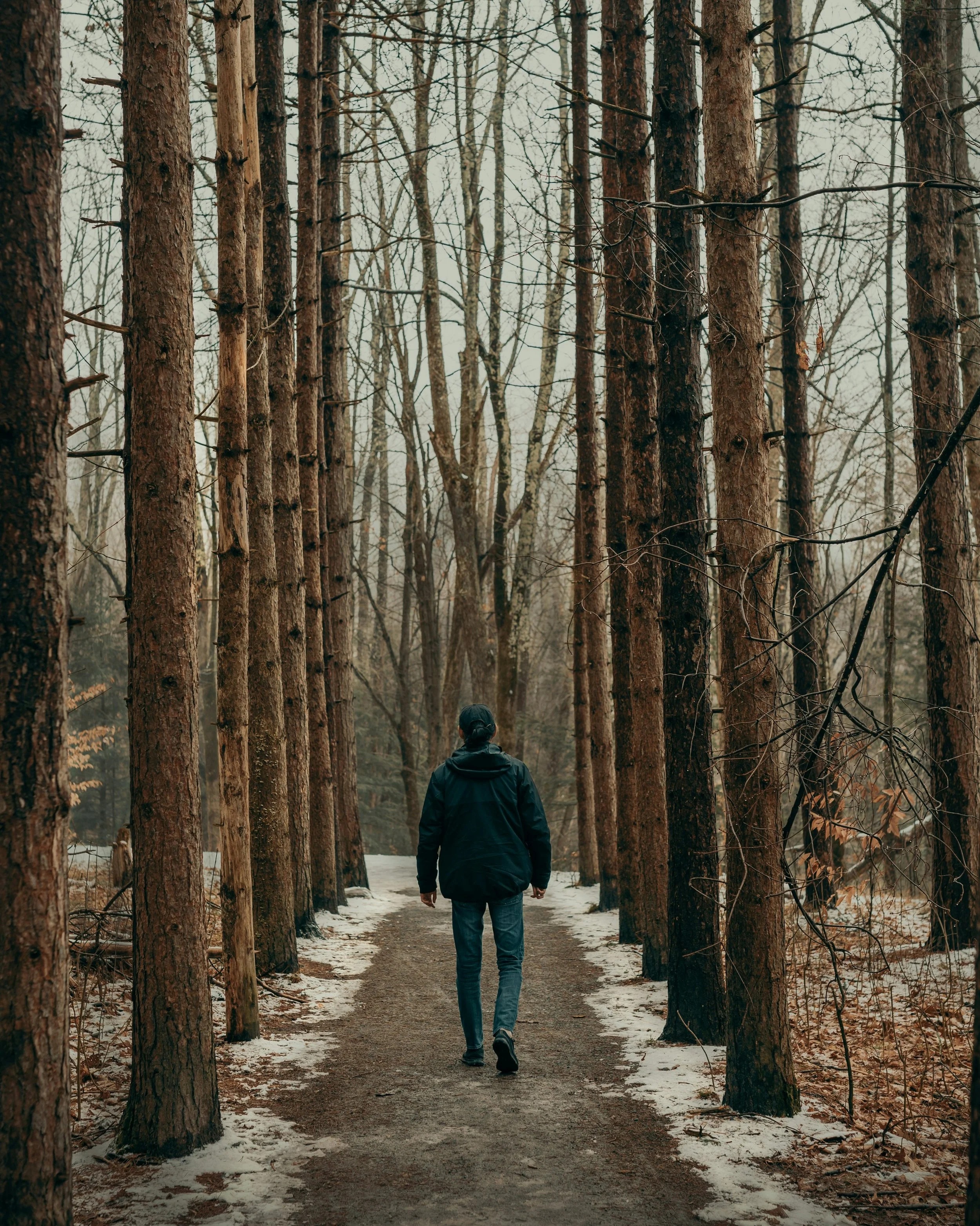 A person walking down a wooded trail with bare trees and snow on the ground during winter.