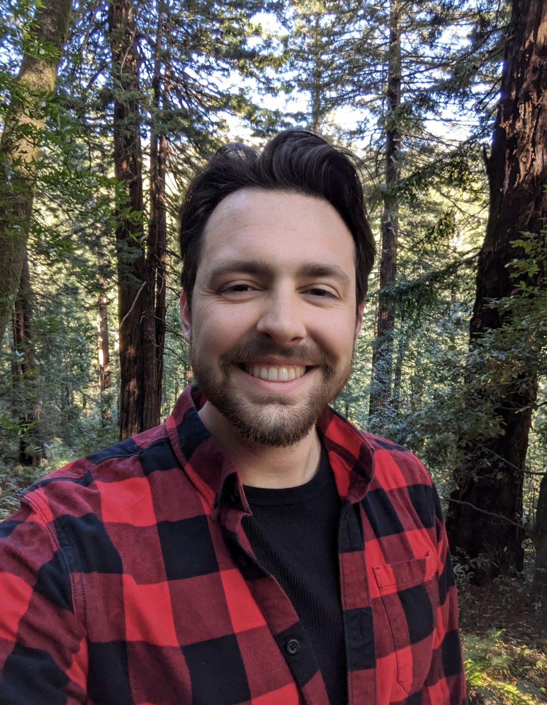 A man taking a selfie in a forest, wearing a red and black checkered shirt, smiling, surrounded by tall trees and green foliage.
