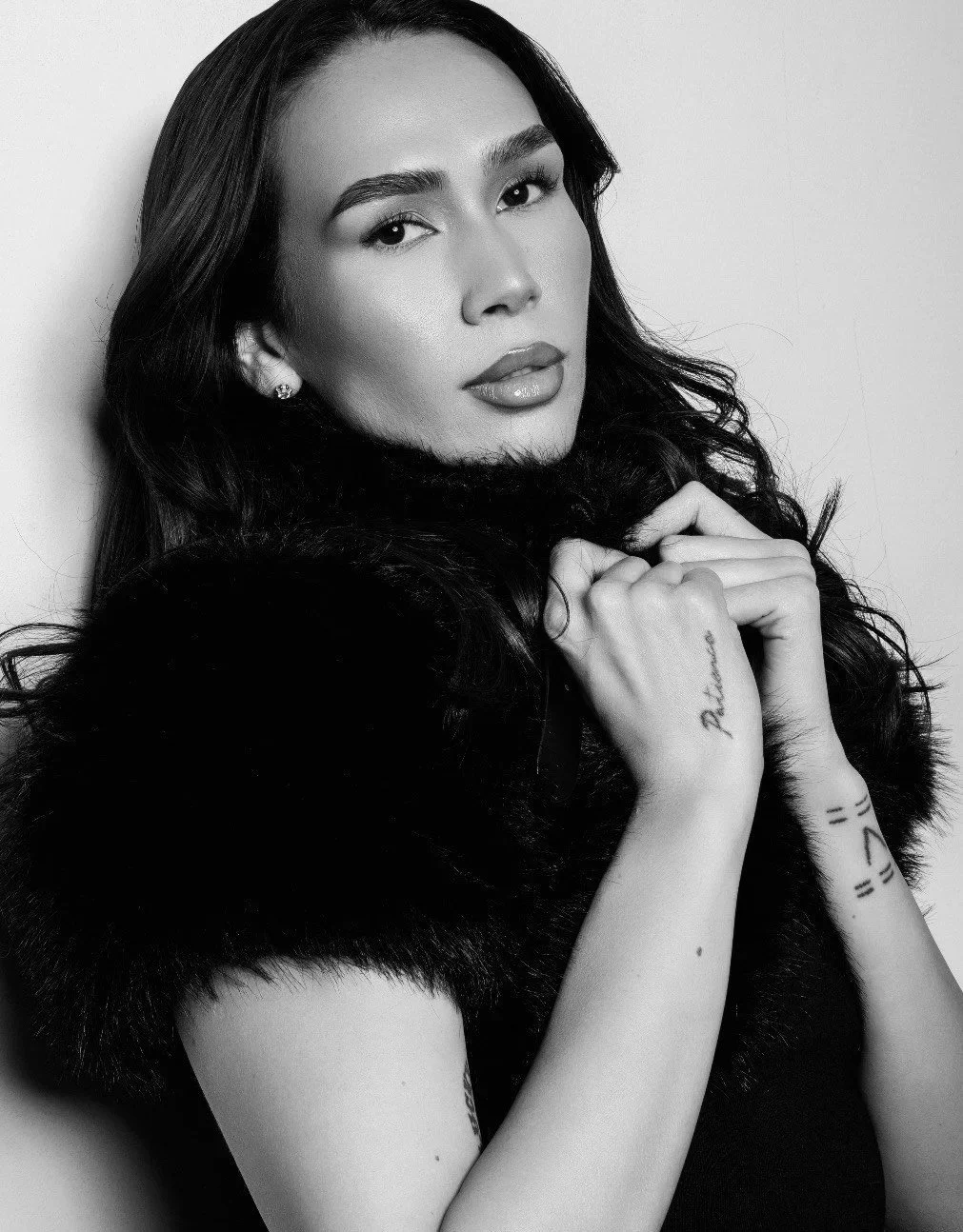 A black-and-white portrait of a woman with long dark hair, wearing a fur coat, looking at the camera with a neutral expression.