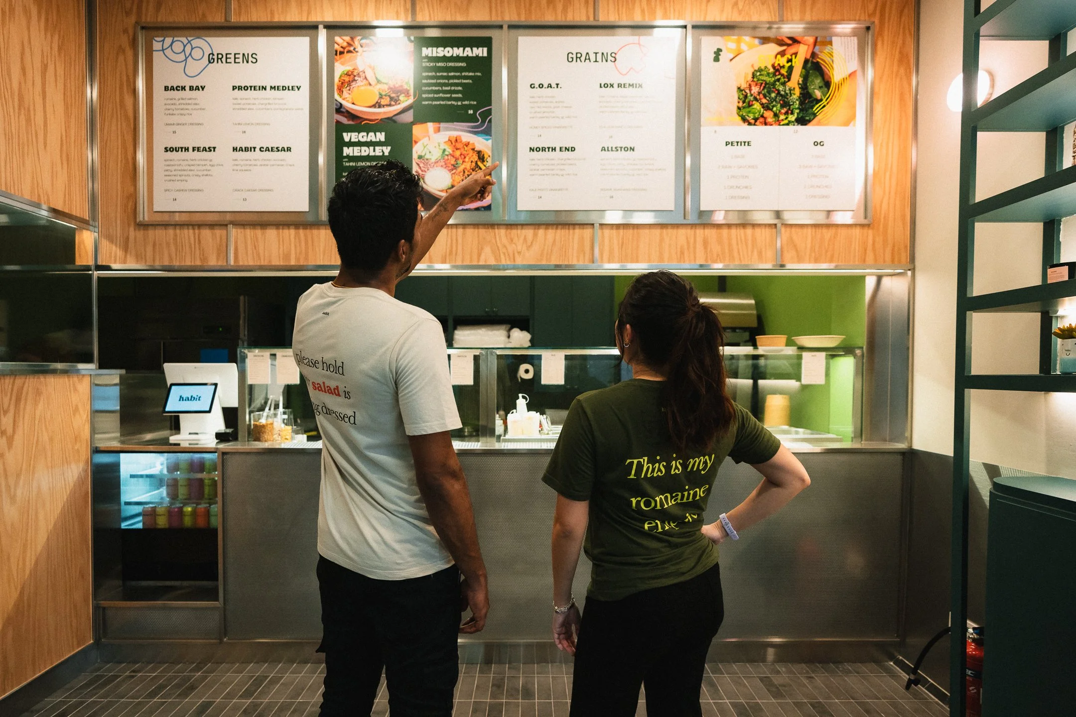 Two people standing at a counter in a restaurant, looking at the menu boards above. One person is pointing at the menu.