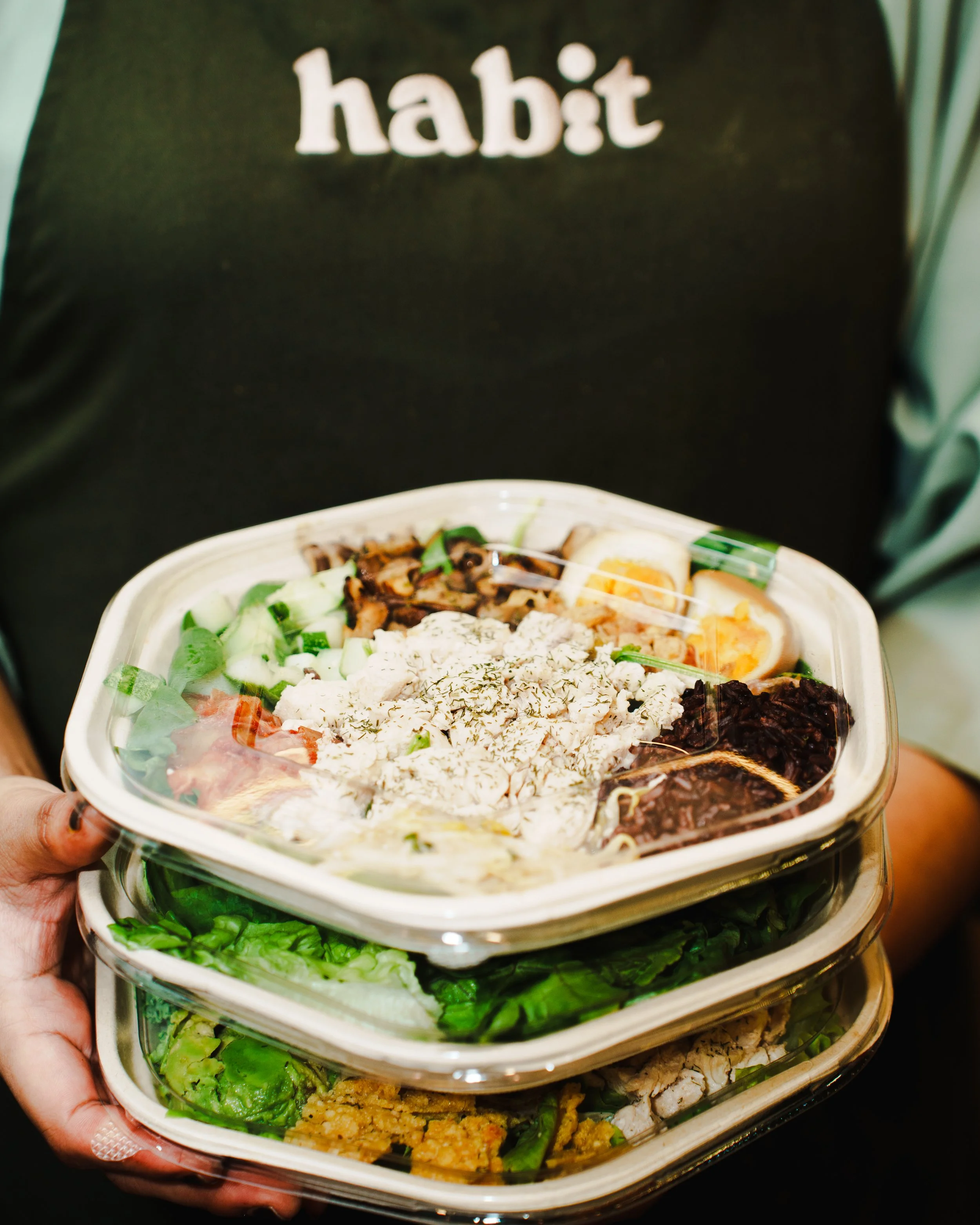 Person holding three stacked takeout containers filled with various salads and toppings, with the word 'habit' visible on a black shirt in the background.
