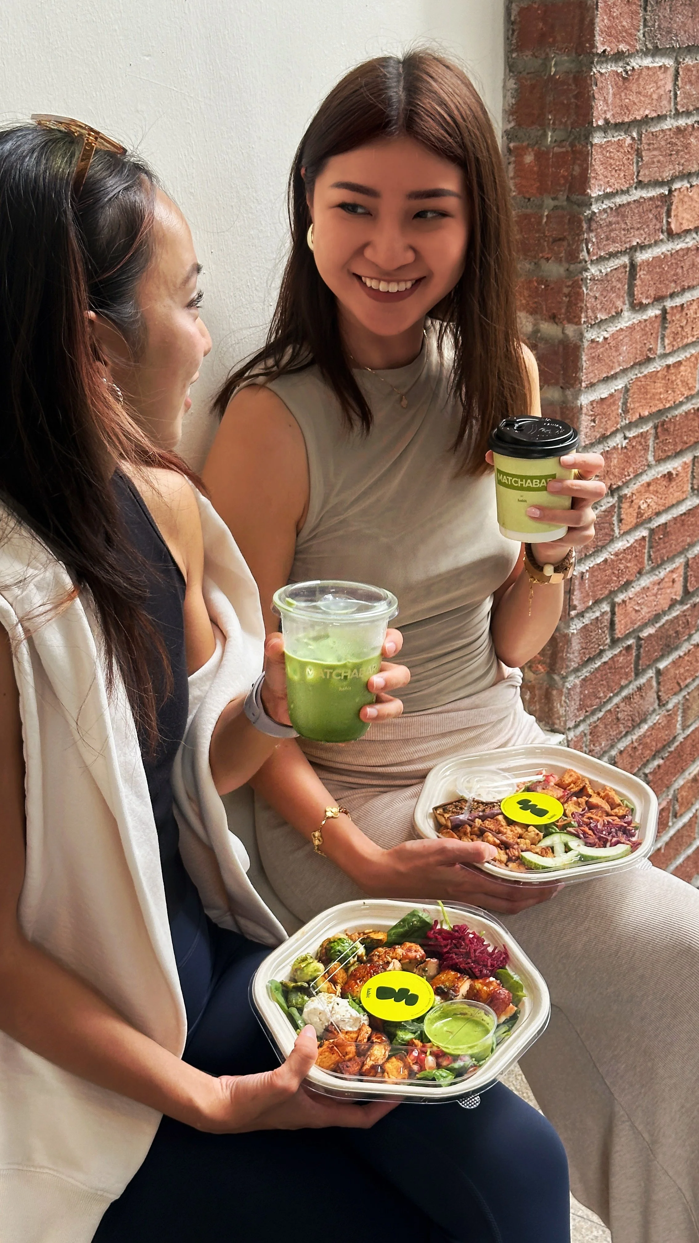 Two women sitting against a brick wall, smiling and holding takeout salads and drinks.