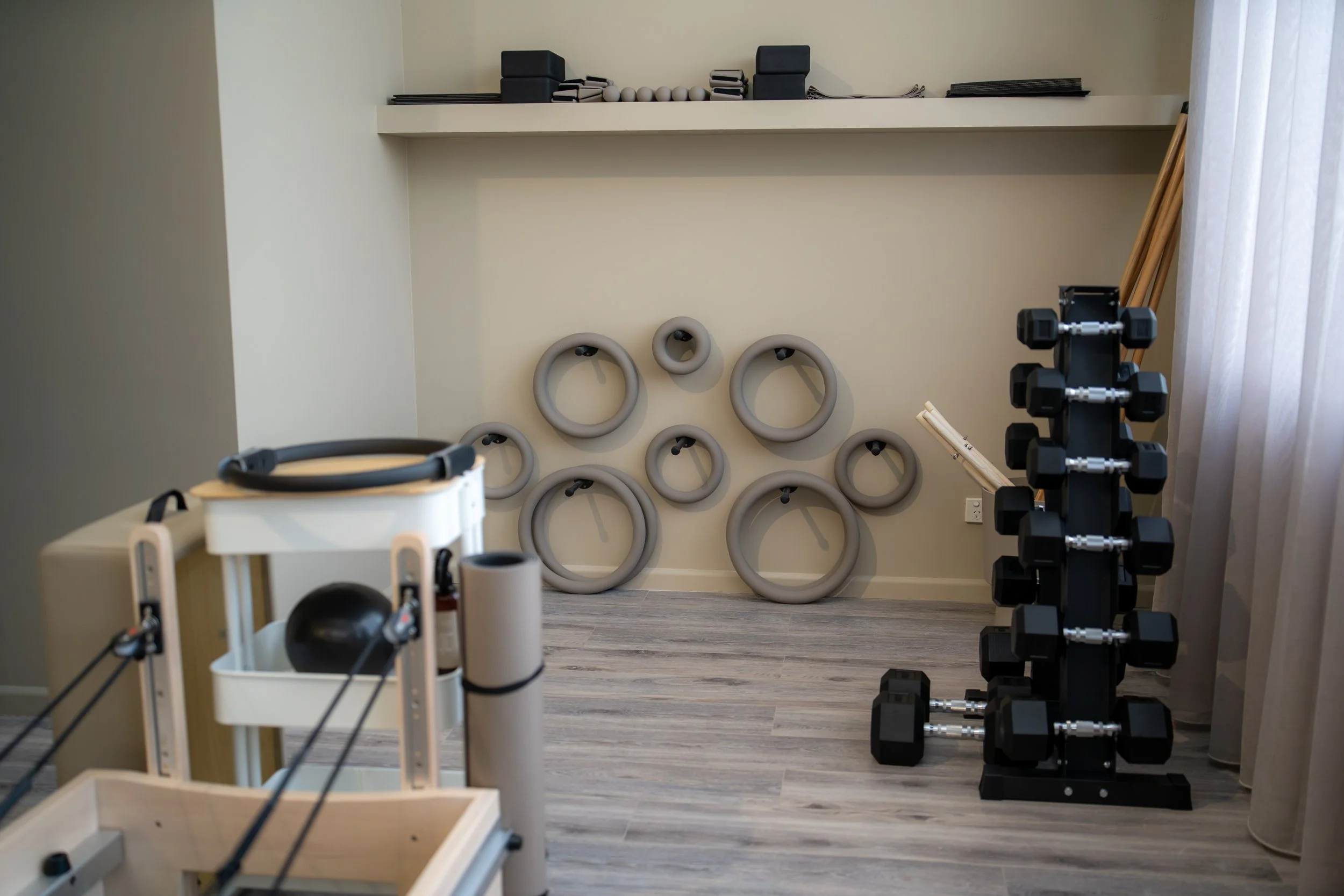 A fitness or rehabilitation room with rubber rings mounted on a beige wall, a black dumbbell rack with various sizes of dumbbells, and a workout machine in the foreground. The room has light wooden flooring, a wooden staircase, and a white curtain on the right side.