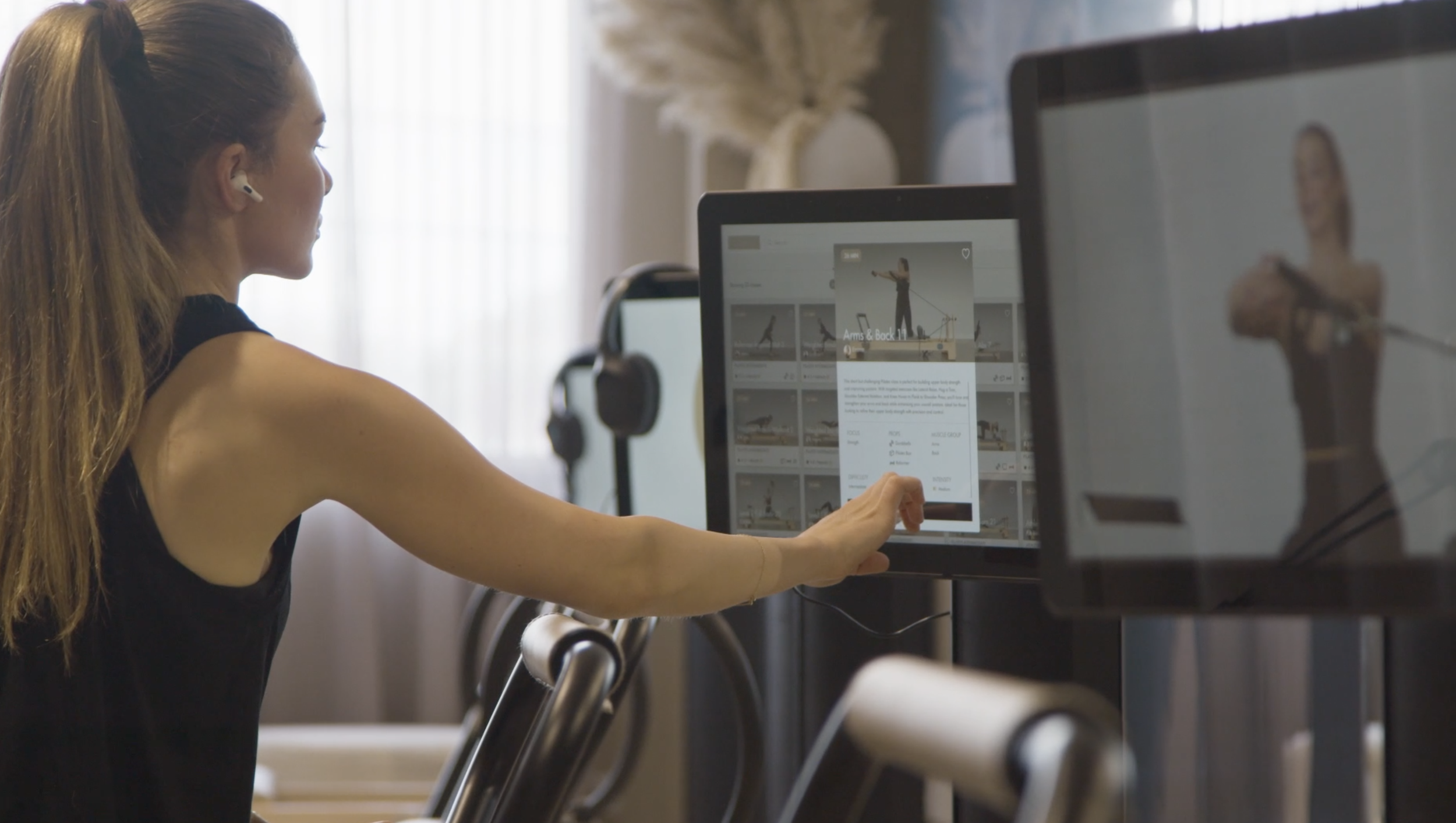 A woman with long hair in a ponytail using a treadmill and touching a mouse in front of a computer monitor displaying workout options.