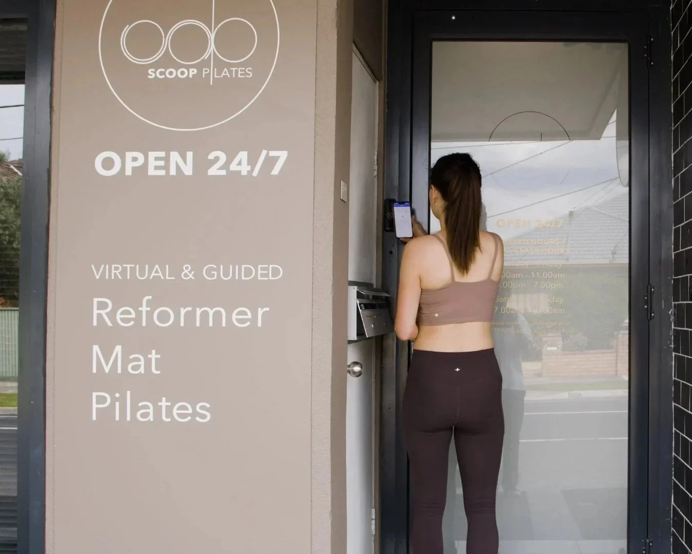 A woman standing at an entrance kiosk of a fitness studio, using her phone to check in. The studio signage indicates it is open 24/7 and offers virtual and guided classes in reformer, mat, and Pilates.
