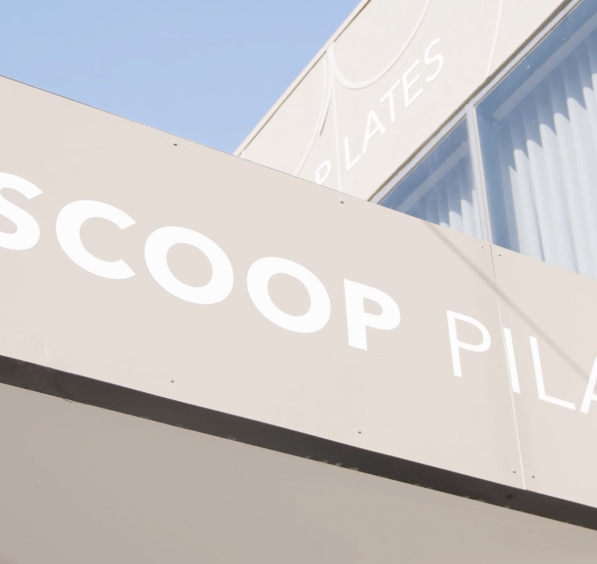 Close-up of a beige building exterior with a partial view of a sign displaying white letters, and a window with blue curtains under a clear blue sky.