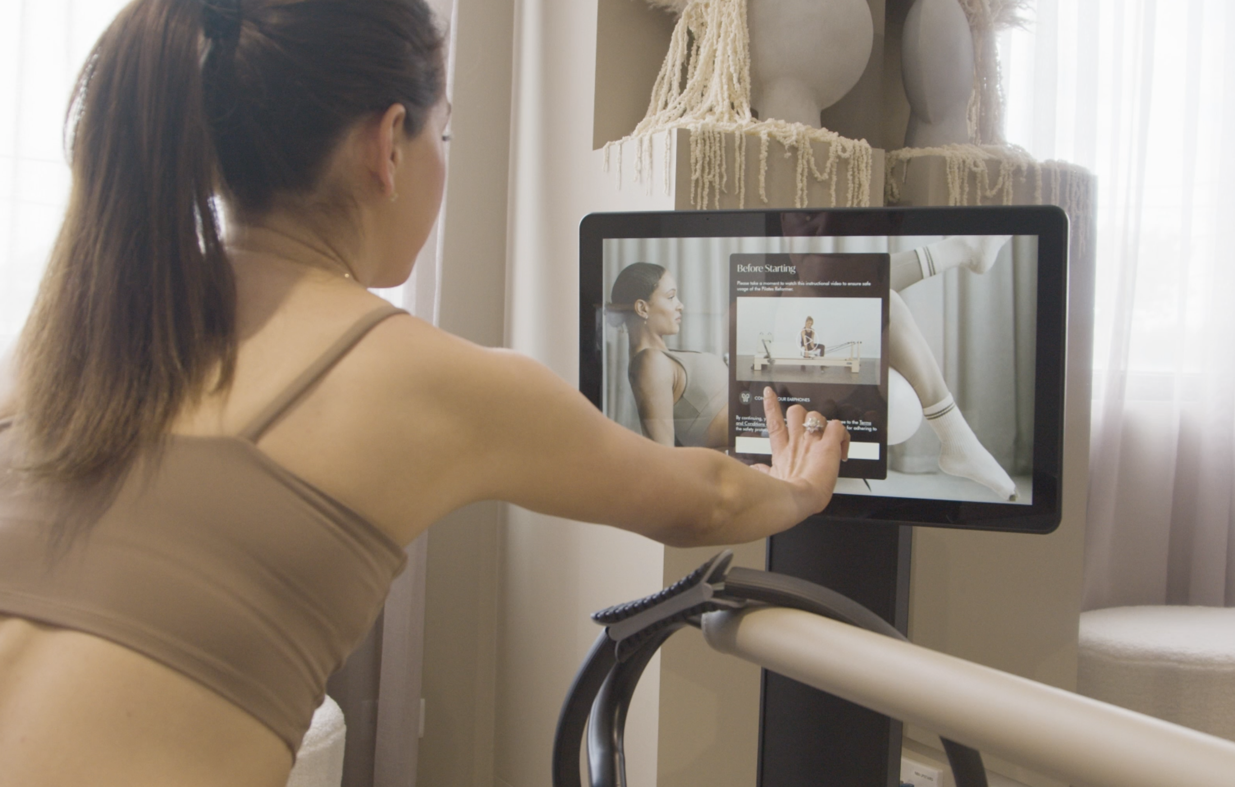 A woman is watching an instructional exercise video on a tablet at a physical therapy or exercise studio. The video shows her profile and a mannequin demonstrating exercises with a resistance band.