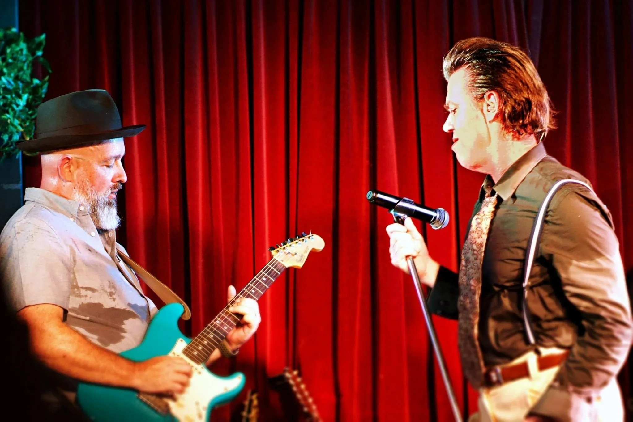A man with a grey beard wearing a hat playing an electric guitar, and a man with slicked hair similar to David Bowie holding a microphone, performing against red curtains.