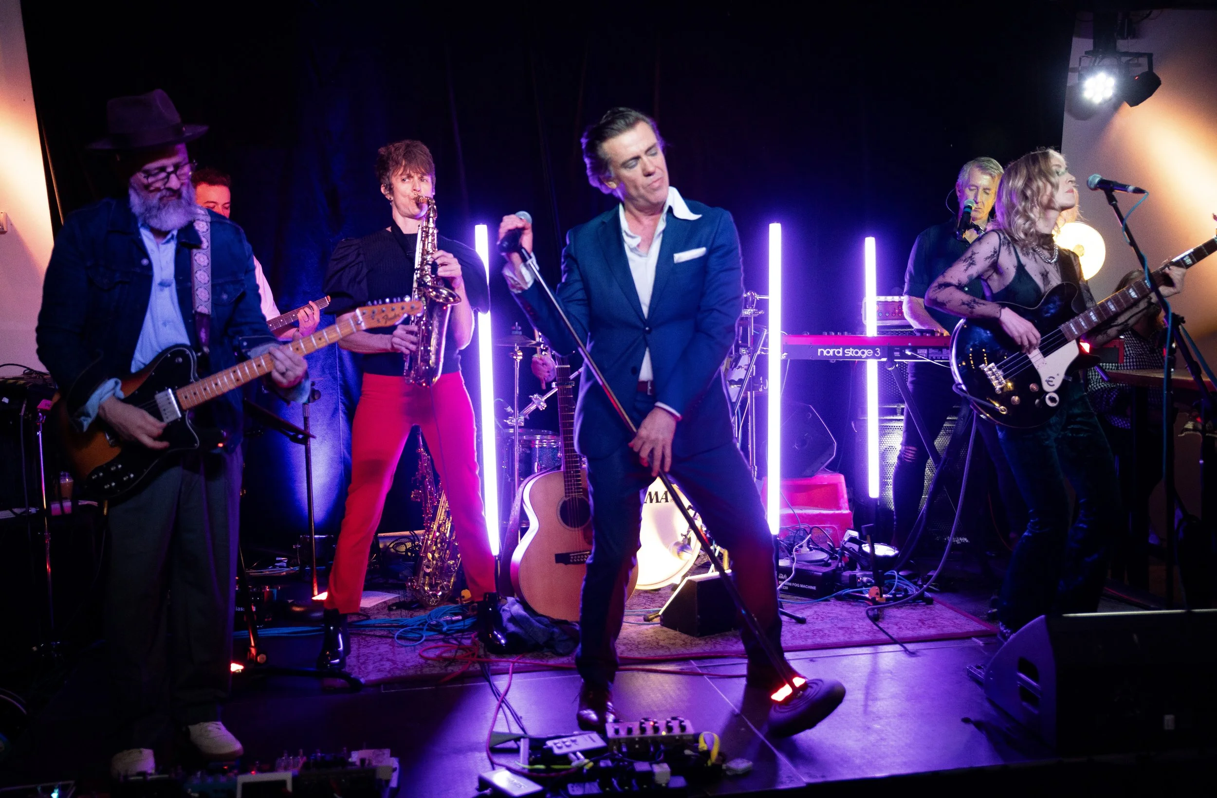Band performing on stage with various instruments including guitars, drums, and keyboard. The stage is lit with colorful lights, and the lead singer is in the center, dressed in a blue suit like David Bowie, making a gesture with their hands.