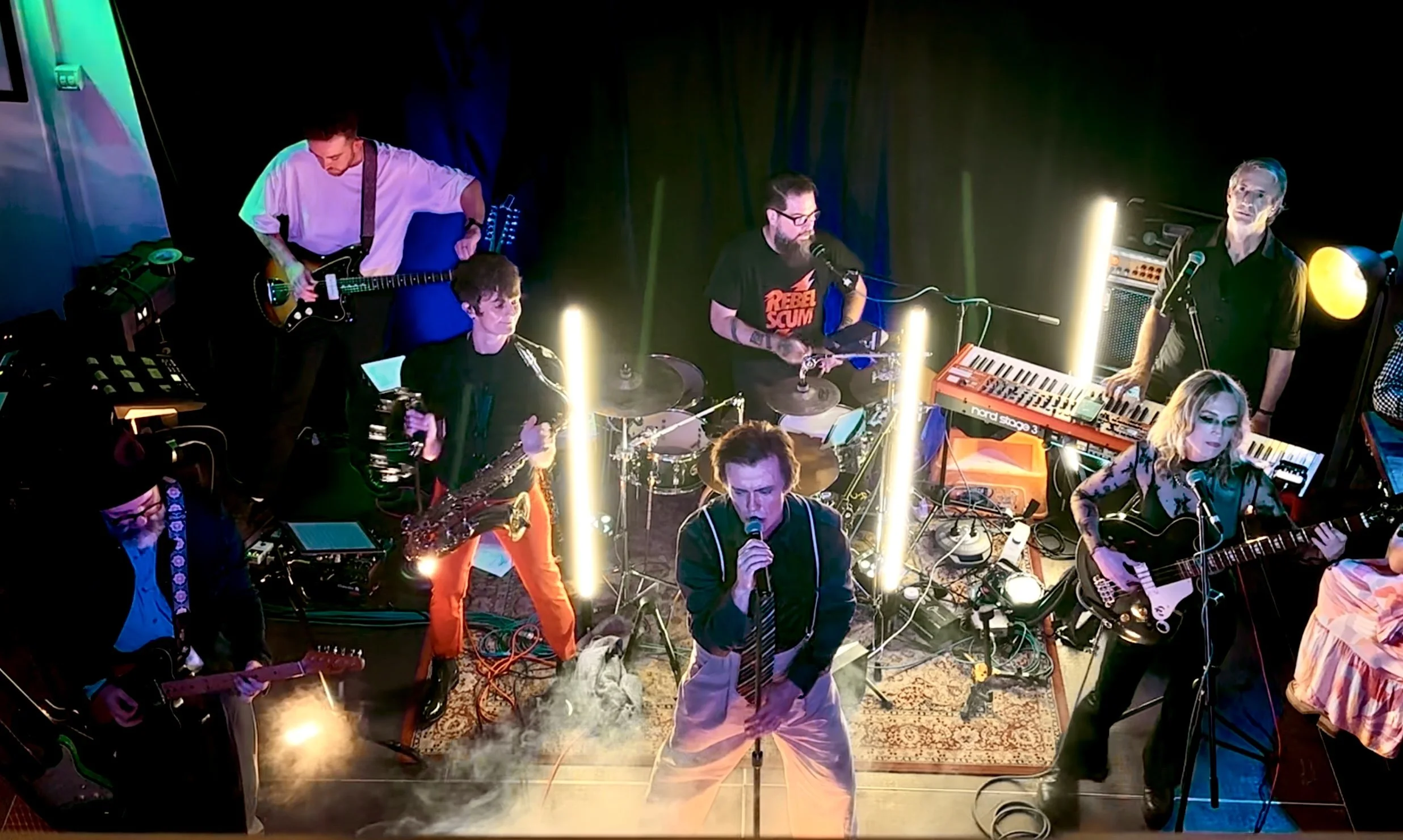 A band performs live on stage with musicians playing various instruments, including guitars, drums, keyboard, and saxophone, under colorful stage lighting.