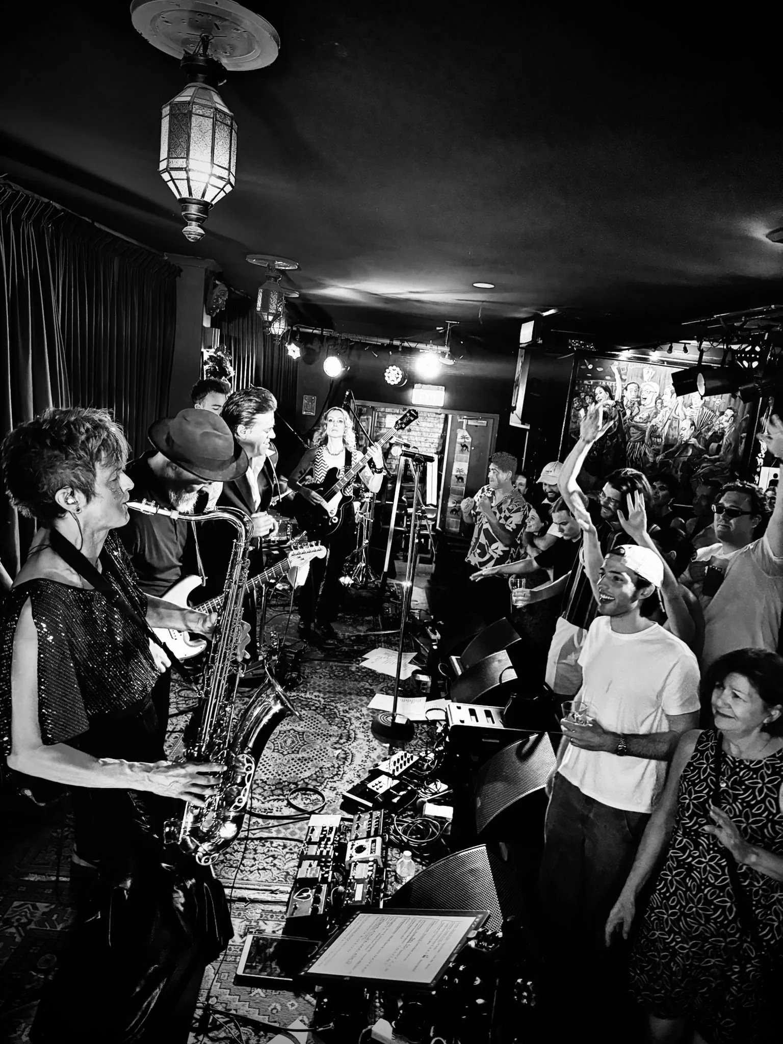 A live music performance in a small venue with musicians playing instruments including guitar and saxophone, and an audience dancing and enjoying the show.
