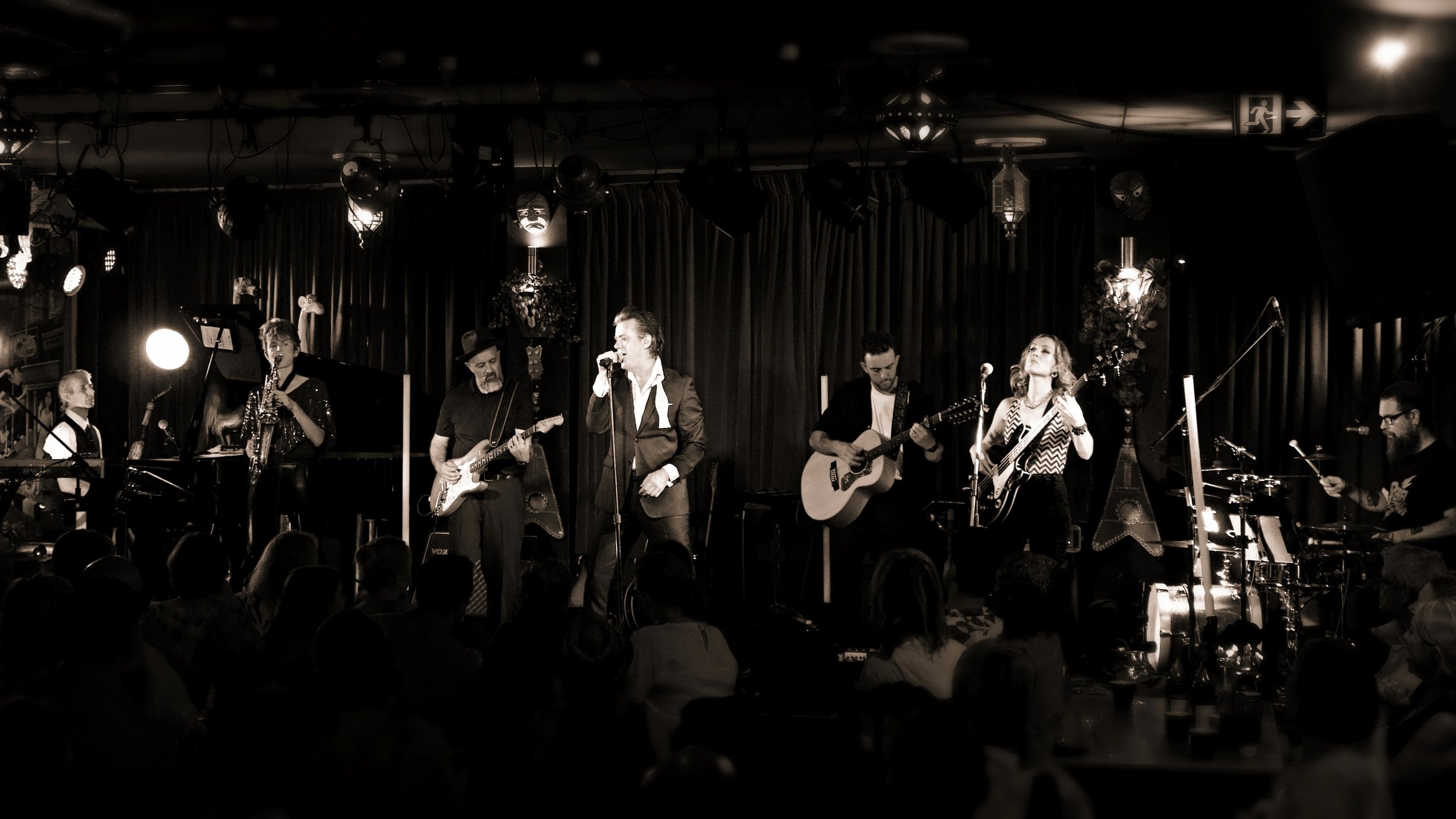 A band performing live on stage with multiple musicians playing instruments and singing, audience visible in front, dark curtains and stage lighting.