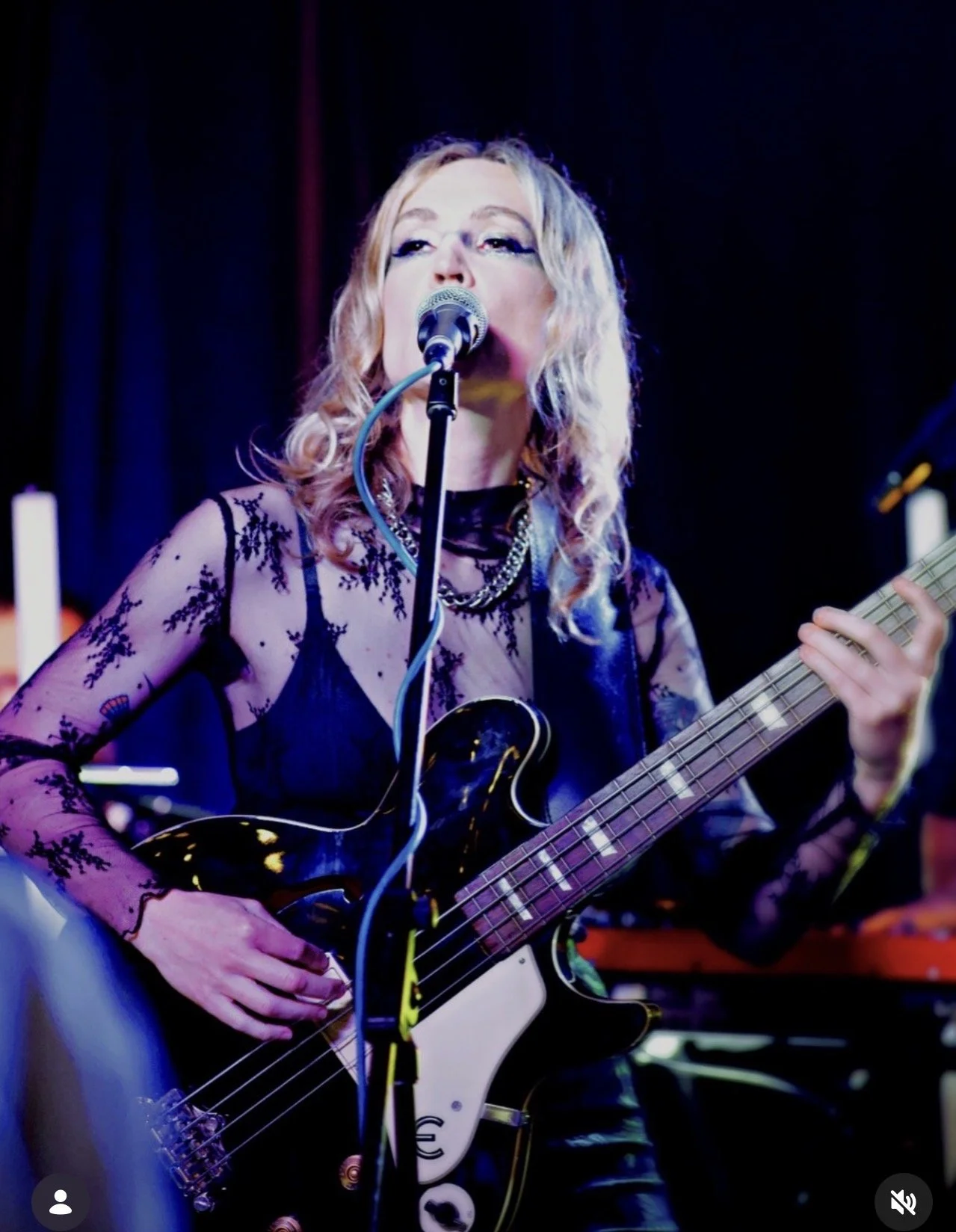 A woman with blonde curly hair singing into a microphone while playing an electric bass guitar, wearing a black lace top and necklaces, on stage with a dark background.