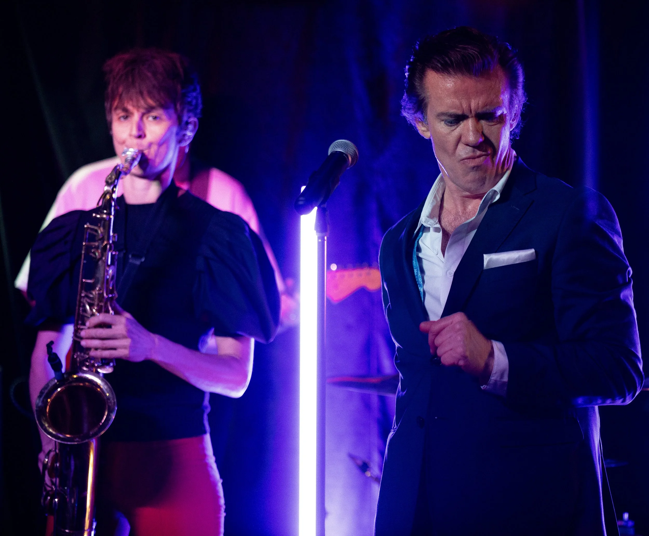 A woman playing the saxophone and a man in a suit with a microphone on stage under colorful lighting.