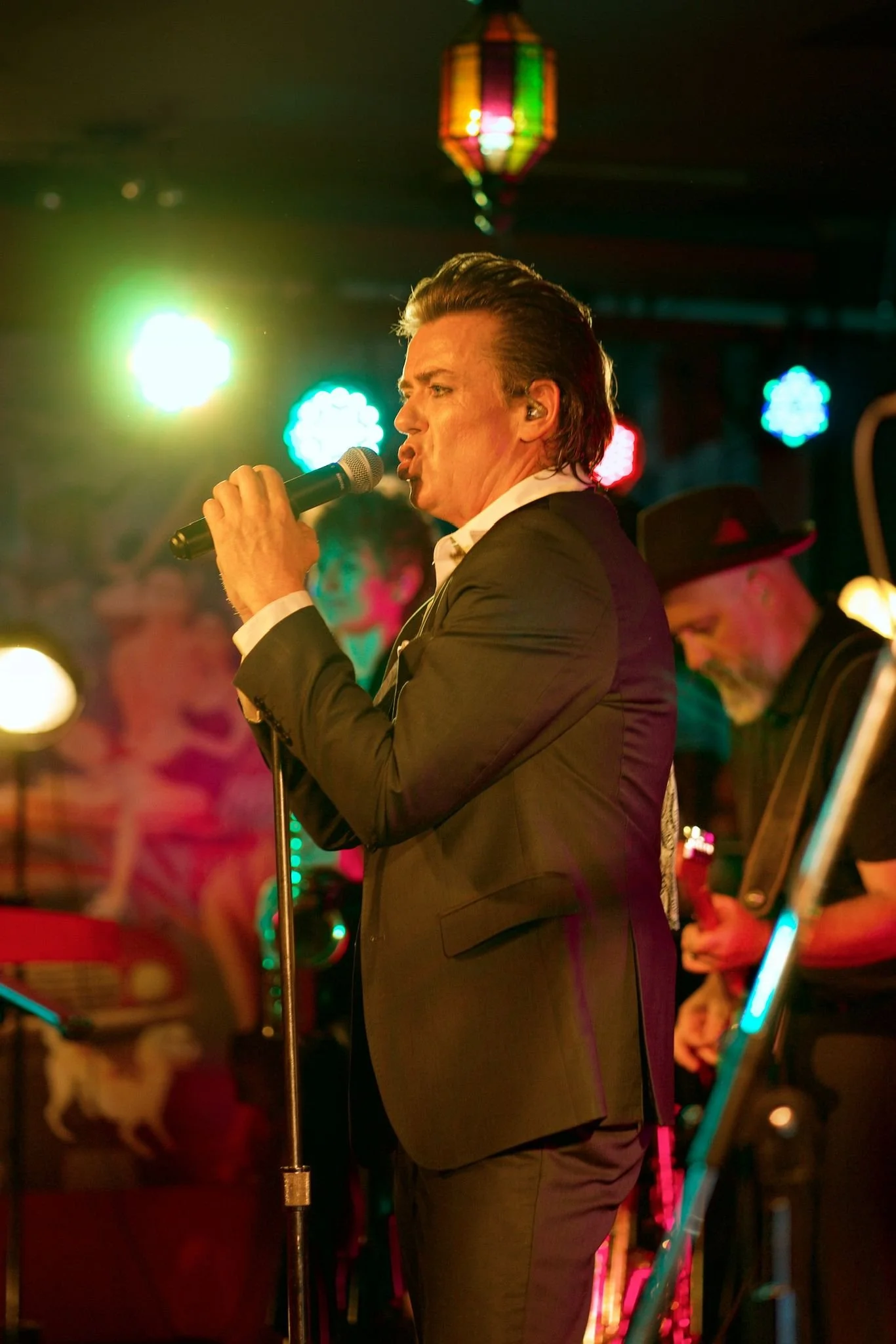 A male singer dressed in a black suit sings into a microphone on stage, with colorful lights and performers in costumes behind him.