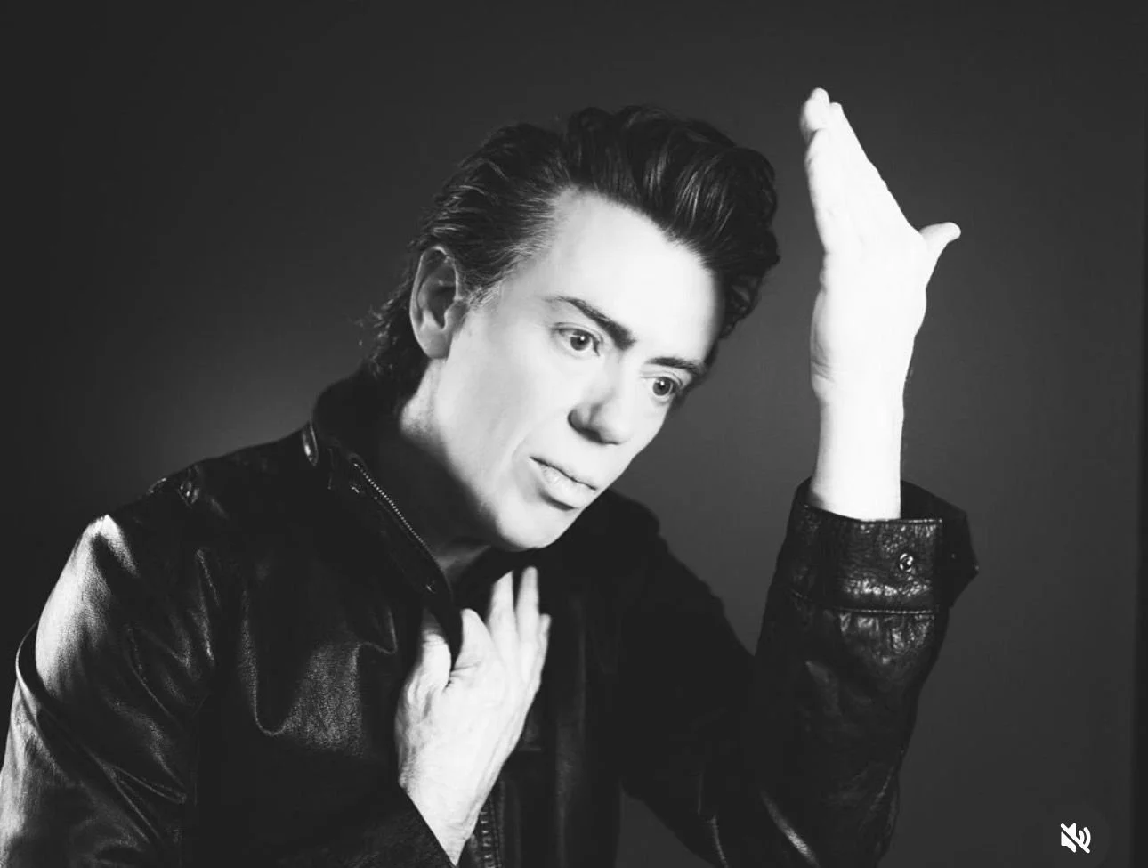 Black and white photo of Ground control singer Dan with slicked back hair wearing a leather jacket, holding one hand to his chest and the other raised with a palm, against a dark background, in the style of David Bowie's Heroes album.