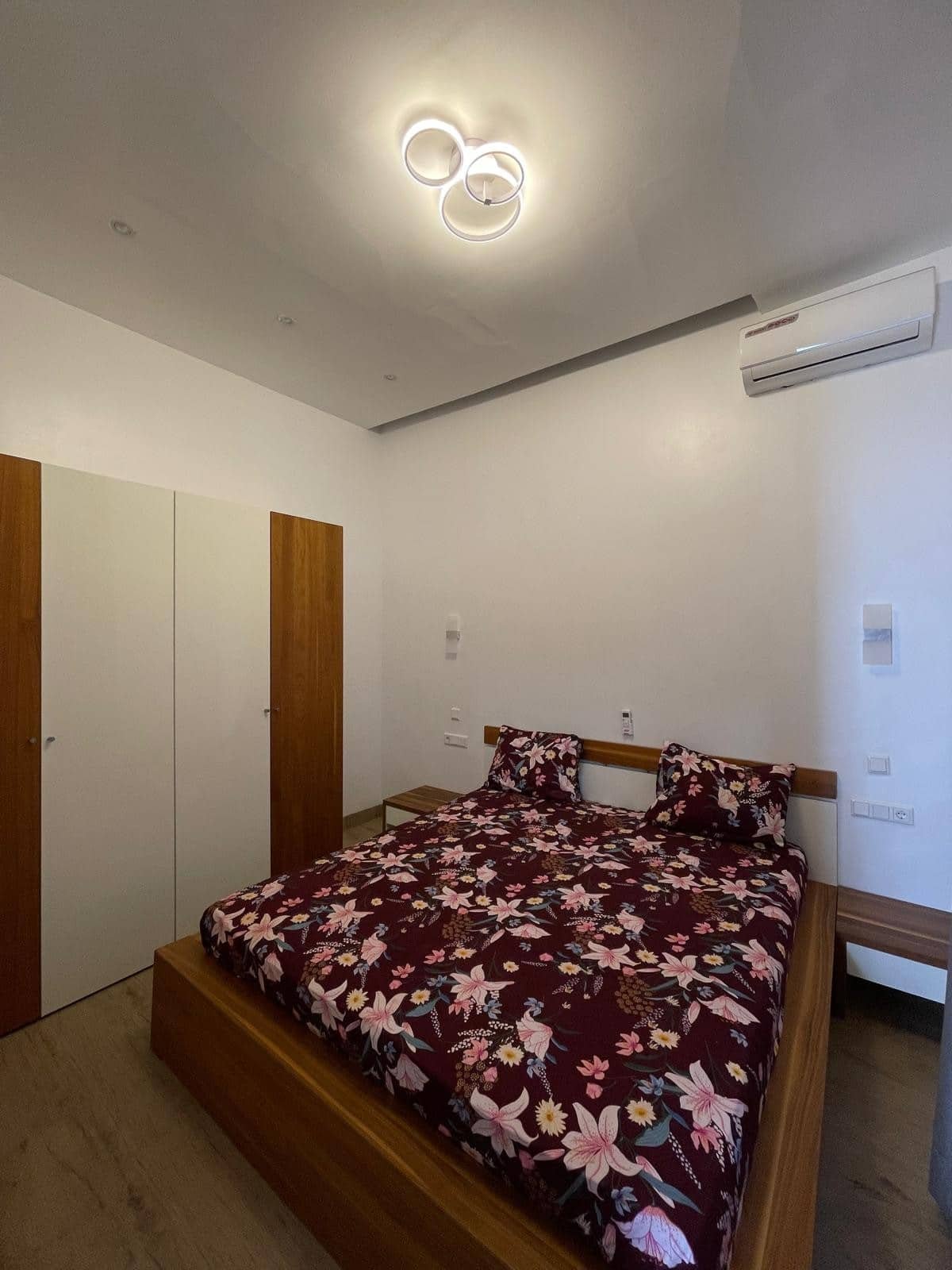 Chambre avec lit en bois, drap à motif floral rouge foncé, deux oreillers, armoire en bois et blanc, table de nuit, climatiseur mural, plafonnier moderne circulaire blancs, murs blancs.