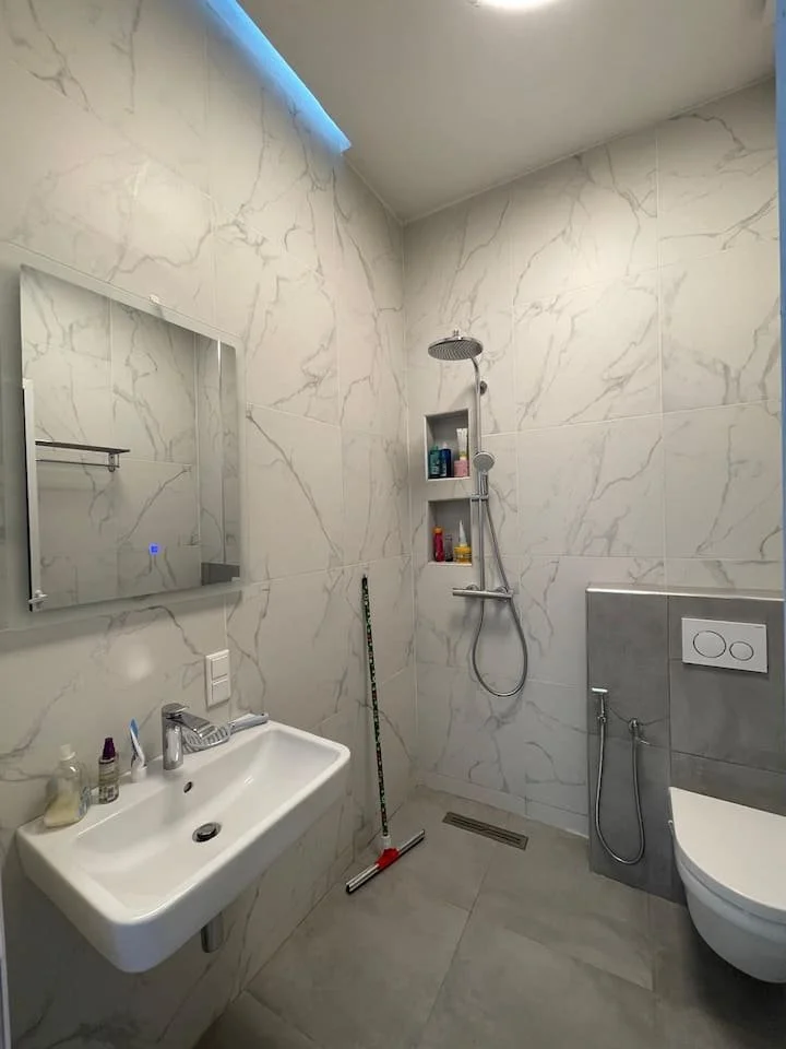 Une salle de bain moderne avec un lavabo blanc, un miroir au-dessus, une douche avec une tête de douche ronde, quelques produits de toilette dans une niche, une éponge et un balai moppant, une toilette suspendue avec un mécanisme invisible, des murs 