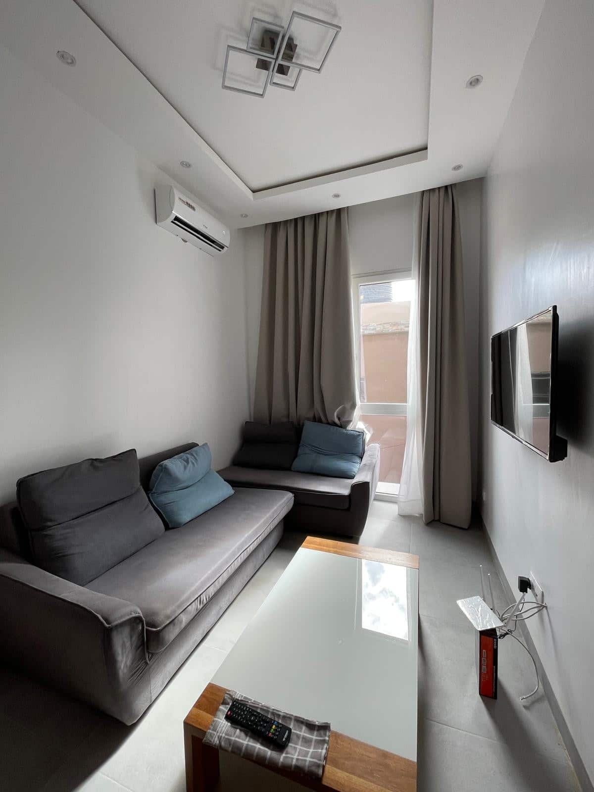 Salon moderne avec canapé gris, coussins bleus, table basse en bois et verre, télévision à l'écran plat, climatiseur, grands rideaux, plafond à plusieurs niveaux avec éclairage encastré, porte-fenêtre avec vue partielle sur l'extérieur