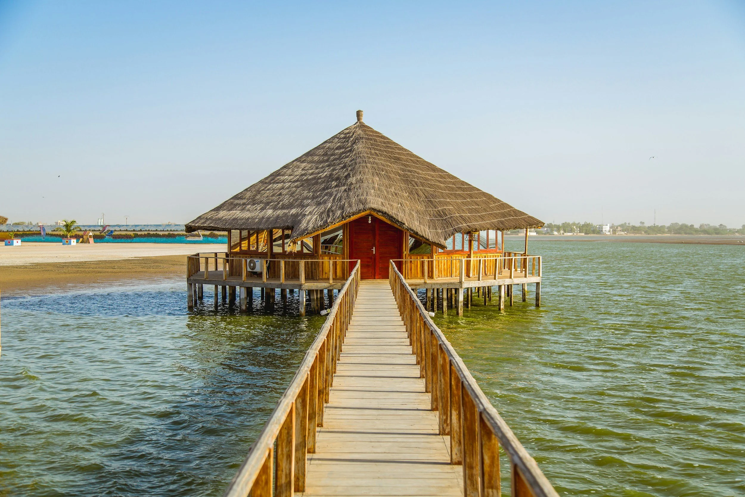 Une cabane en bois construite sur pilotis dans l'eau, accessible par un pont en bois. La cabane a un toit en chaume et une terrasse en bois entourée d'une rambarde.