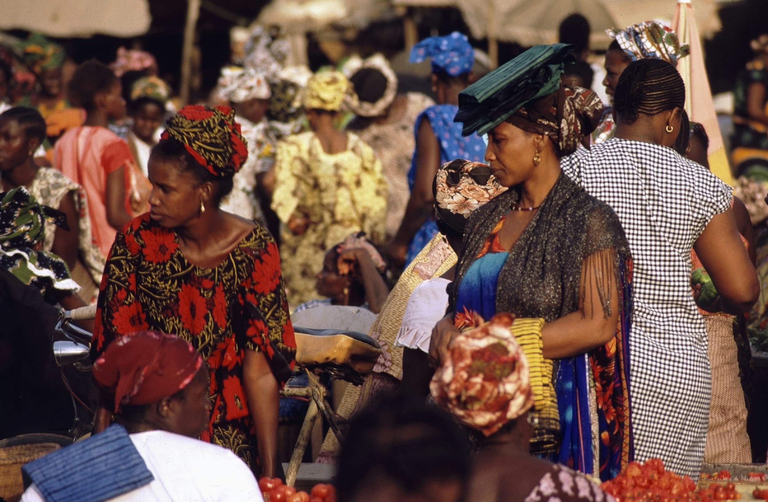 Femmes et jeunes filles africaines portant des vêtements traditionnels colorés, portant des turbans ou des chapeaux, dans un marché en plein air.