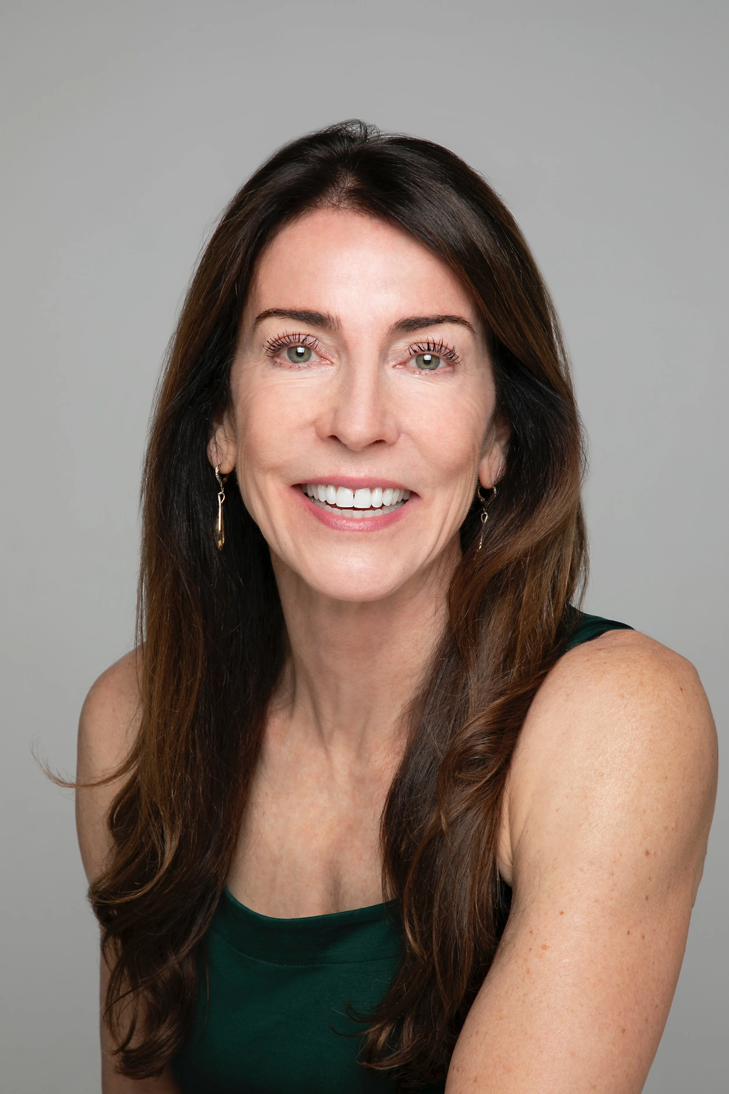 A woman with long, wavy brown hair, green eyes, and earrings, smiling while wearing a dark green top, against a grey background.