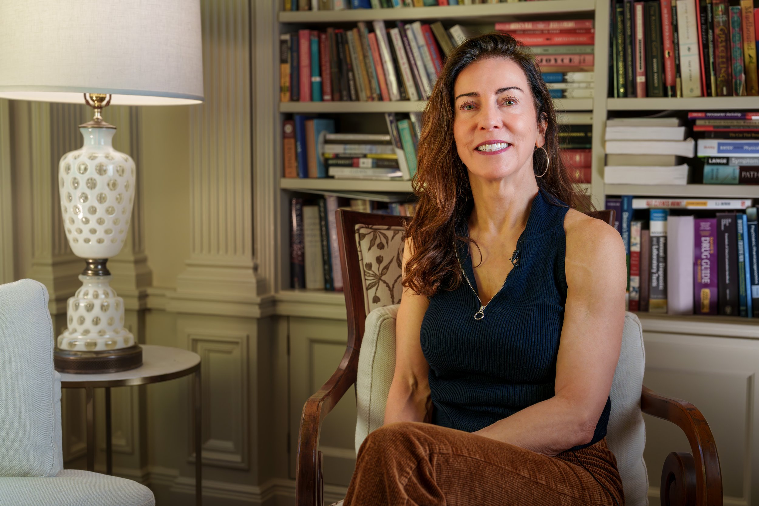 A woman with long, wavy brown hair, wearing a sleeveless black top, sitting on a chair in a room with a bookshelf filled with books in the background, and a table lamp on a round table next to her.
