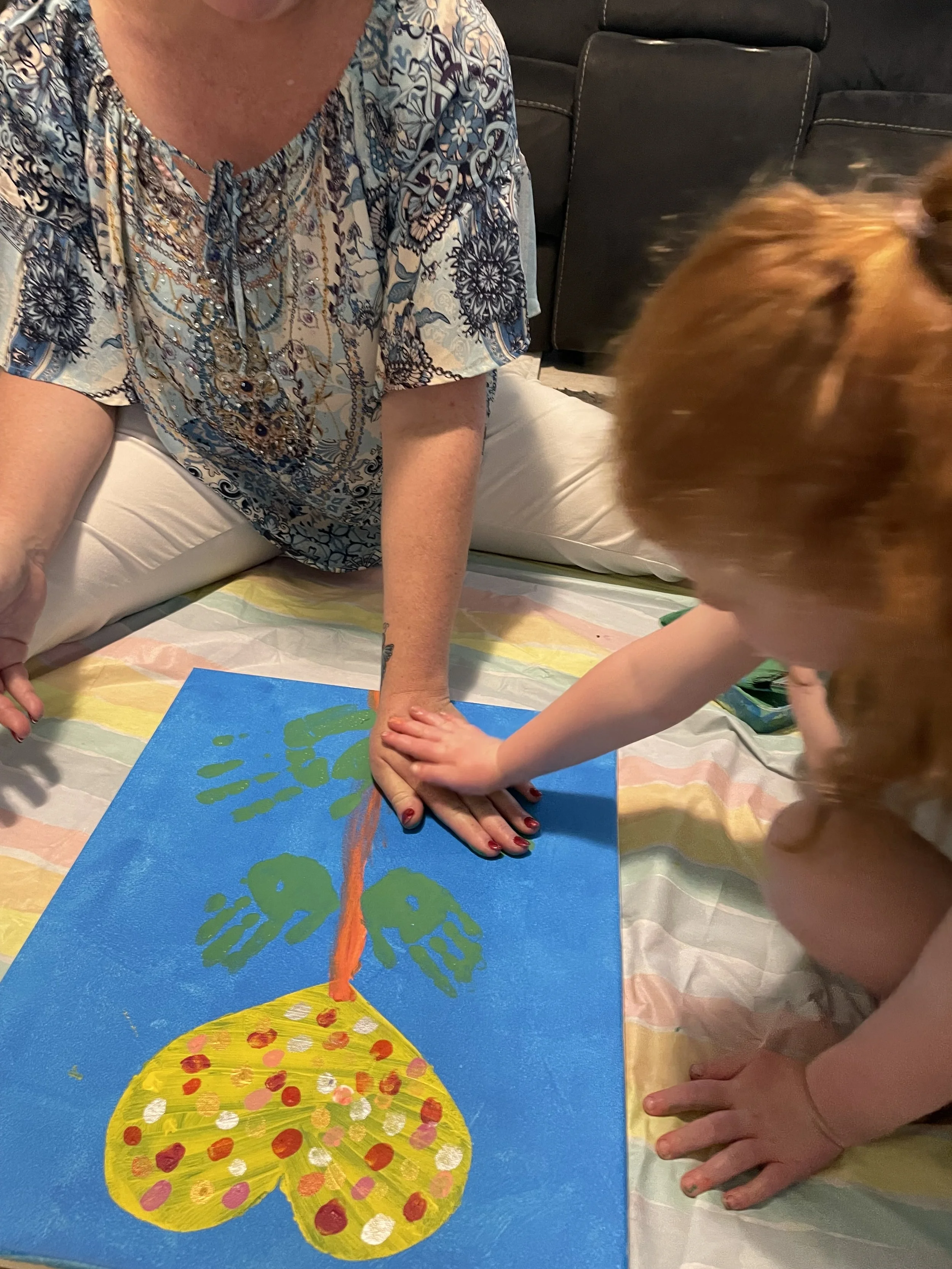 child's hand pressing on adult hand to apply paint to a canvas with handprints making the shape of a flower