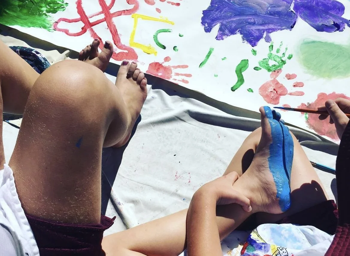 young person painting their foot blue in front of a canvas with paintings on it. Beside them is another young person's feet.