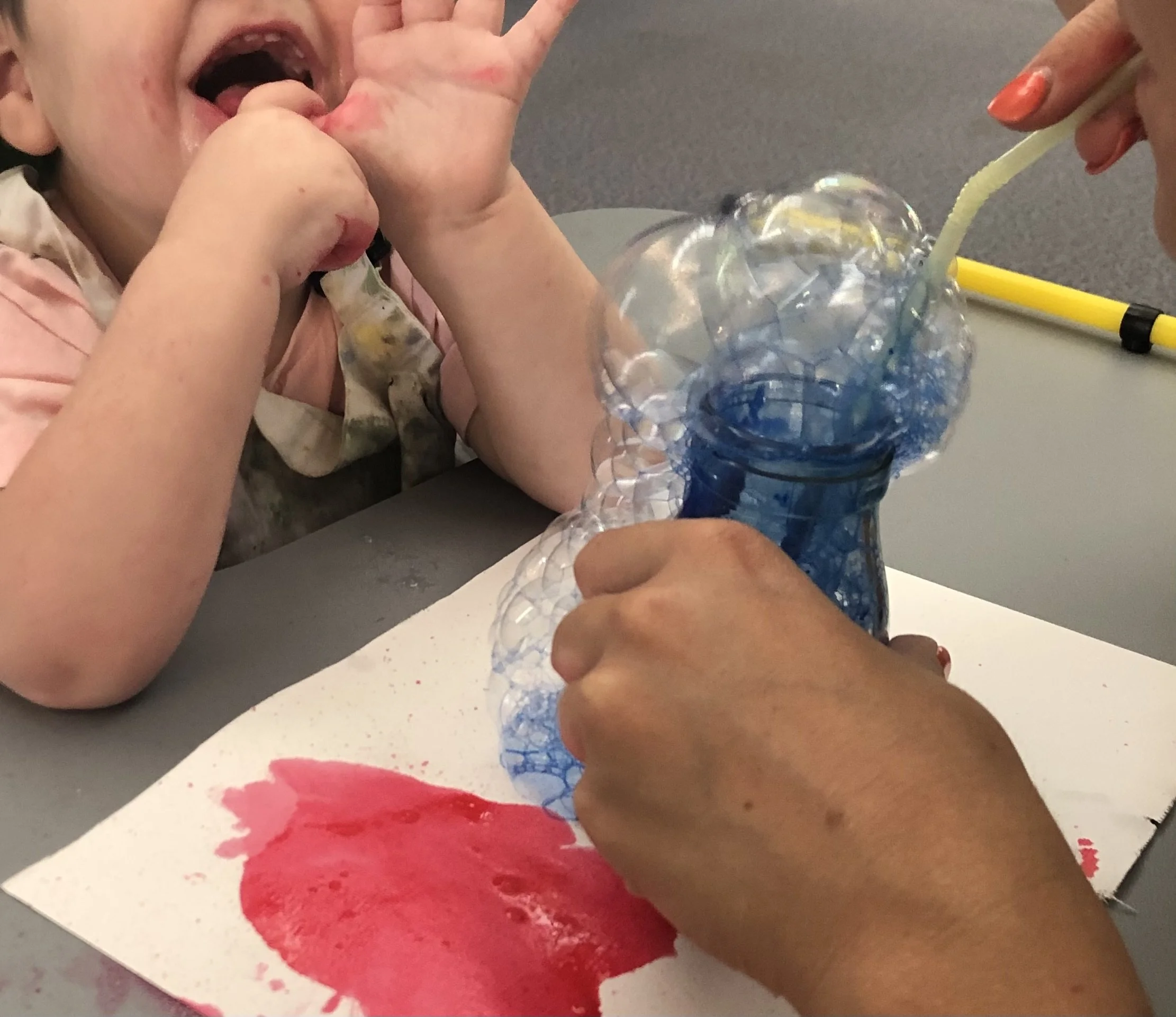 hand holding glass jar filled with blue bubbles onto paper with a red paint mark. In background is the hands and bottom part of face of a child laughing