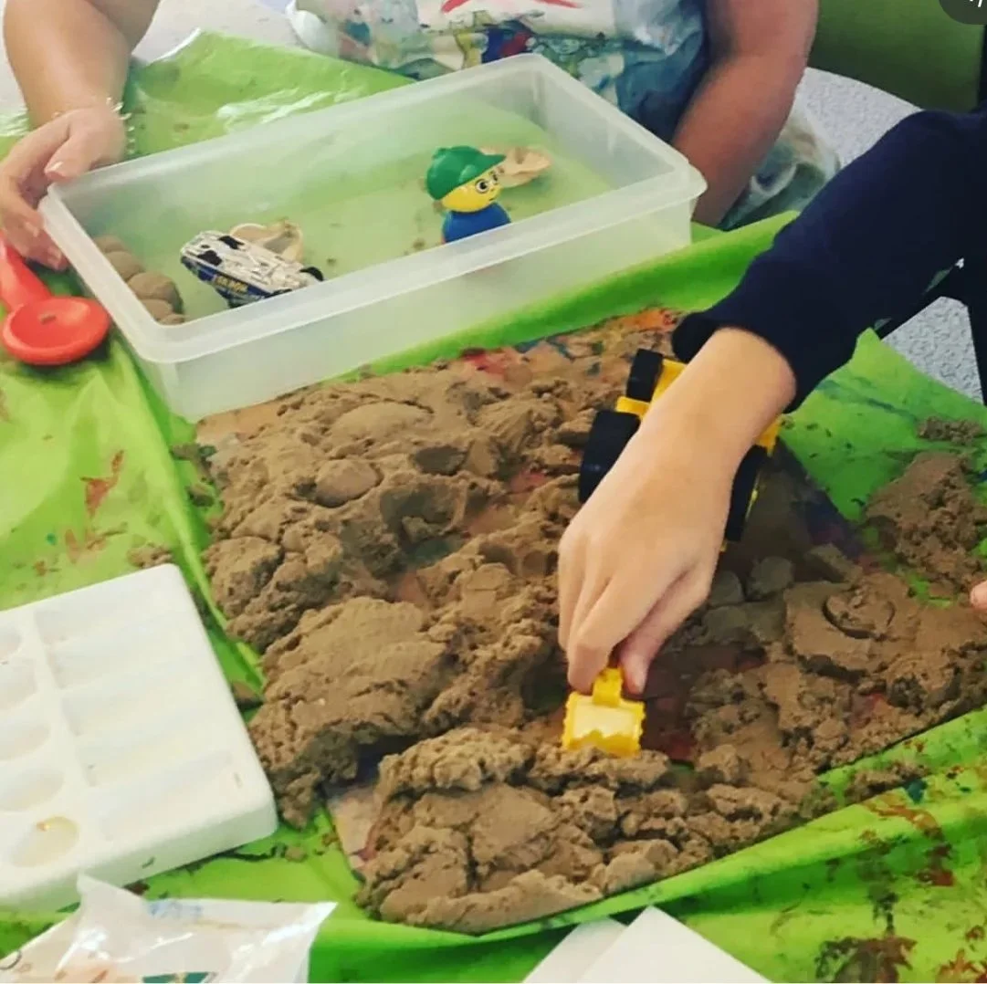 sand on table with young person's hand reaching in with a toy truck. Beside sand is a container with more toys