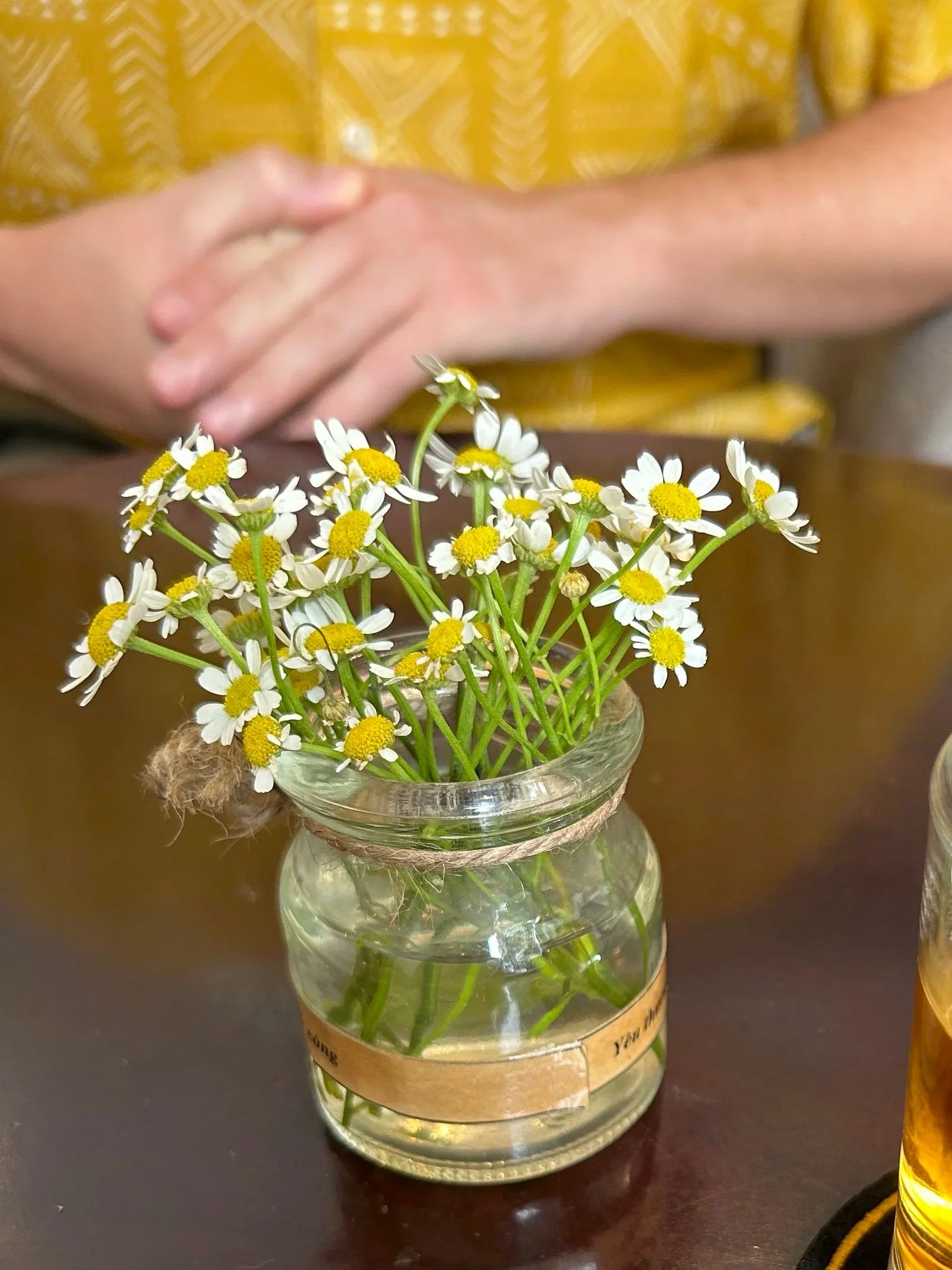 class jar with chamomile flowers on table. In background is a male but you only see his hands clasped in front of him