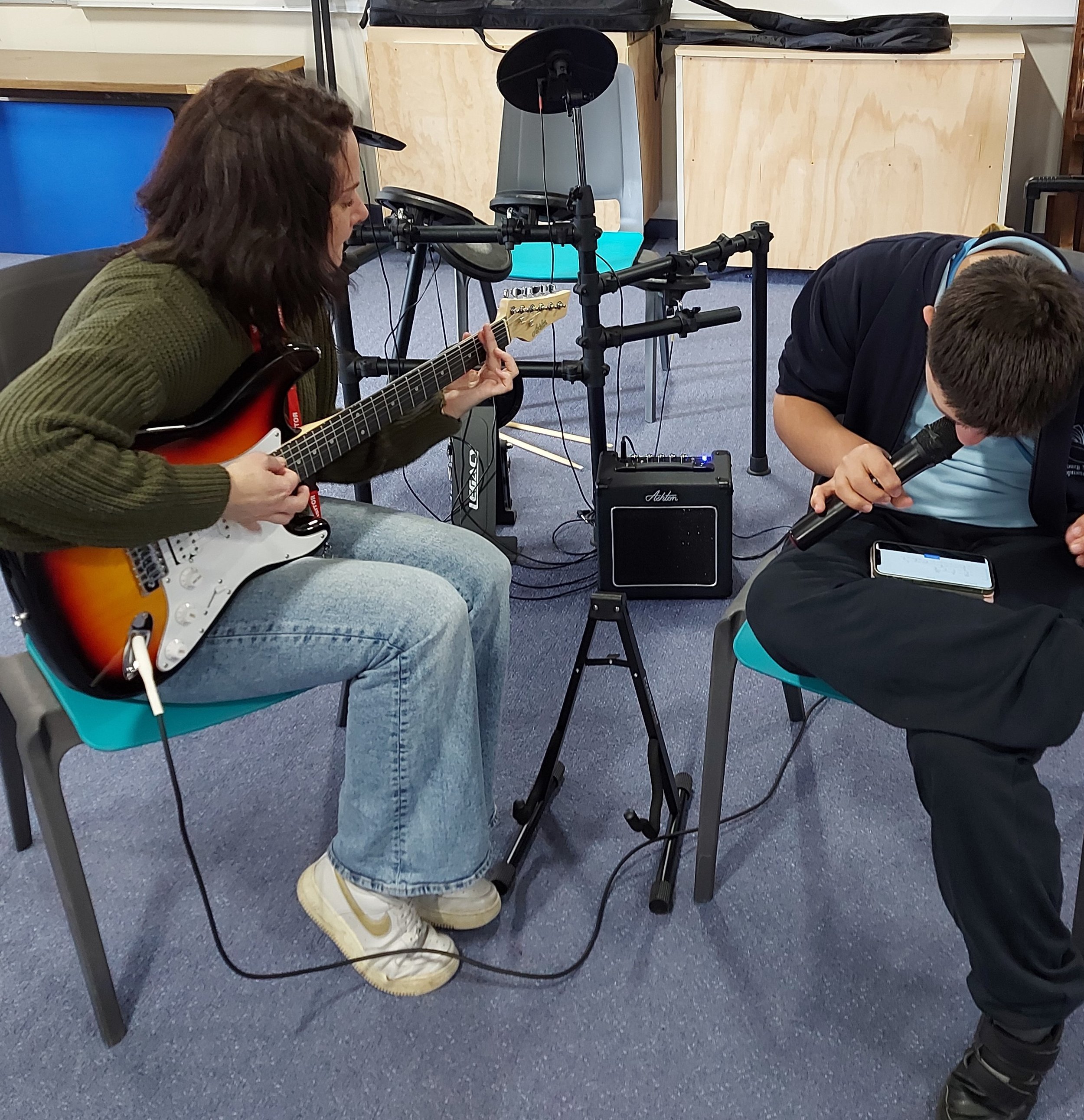 female sitting playing electric guitar with young person in school uniform sitting beside on them, head down singing in to microphone