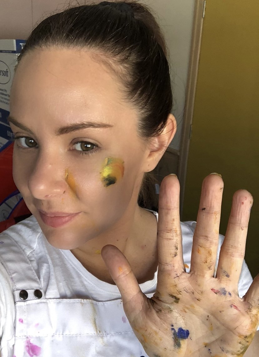 female person with paint on face, smiling at camera with raised hand to say hello