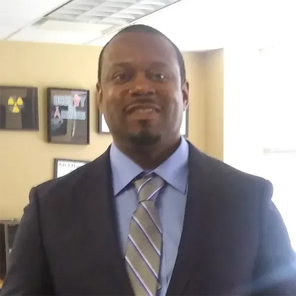 A man in a suit and tie smiling indoors, with framed pictures on the wall behind him.