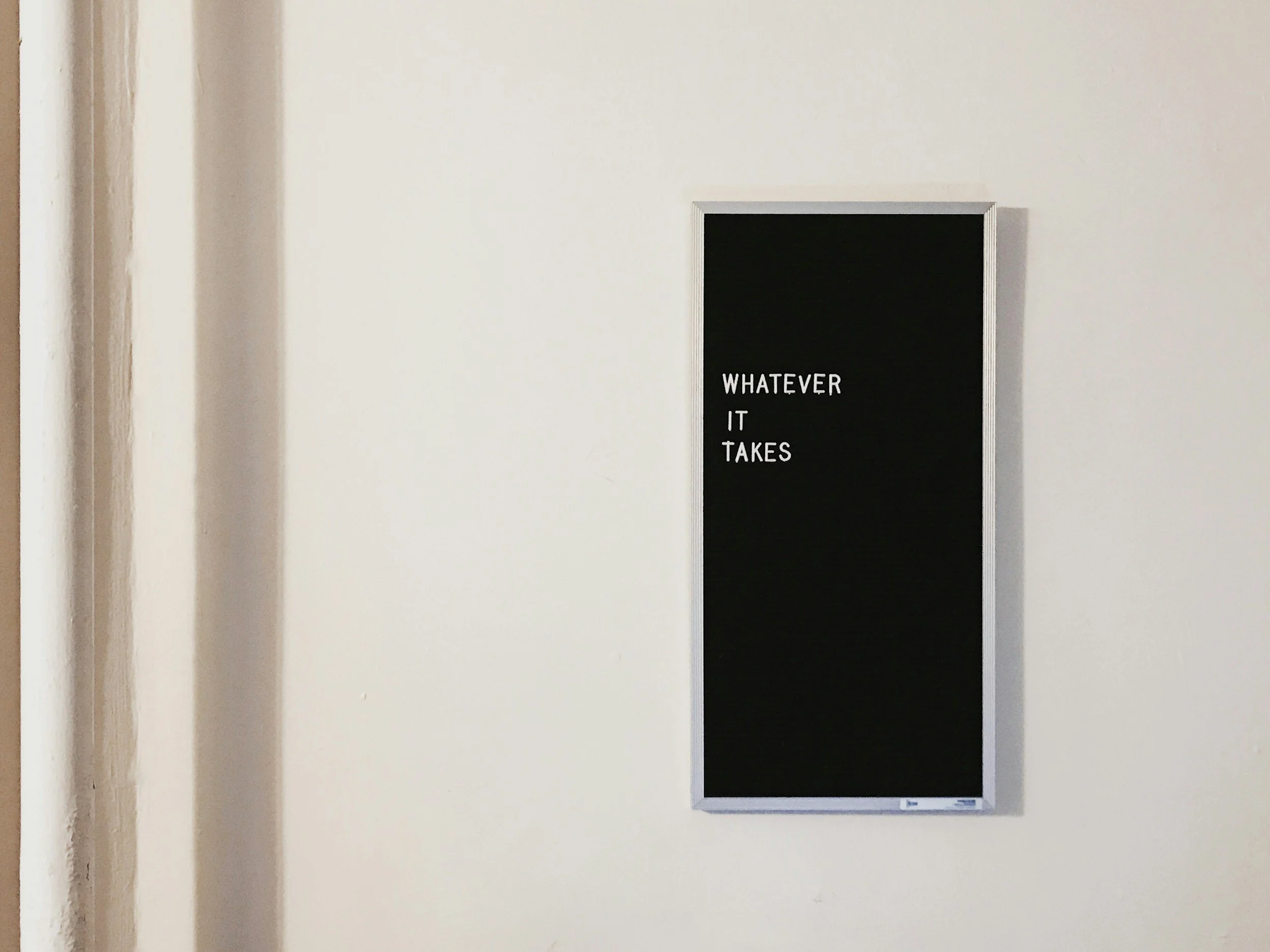 A black wall-mounted message board with a silver frame displaying the text "WHATEVER IT TAKES" on a white wall.