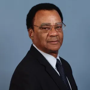 Portrait of a middle-aged man wearing glasses, a dark suit, white shirt, and dark tie against a light blue background.