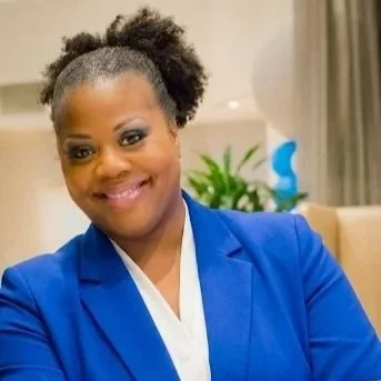 A woman smiling in a blue blazer in an indoor setting with a plant and curtains in the background.