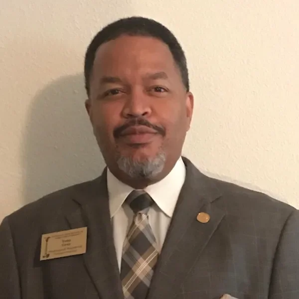A man with short hair and a mustache wearing a checkered tie and a suit with a name tag and pin, standing against a plain wall.