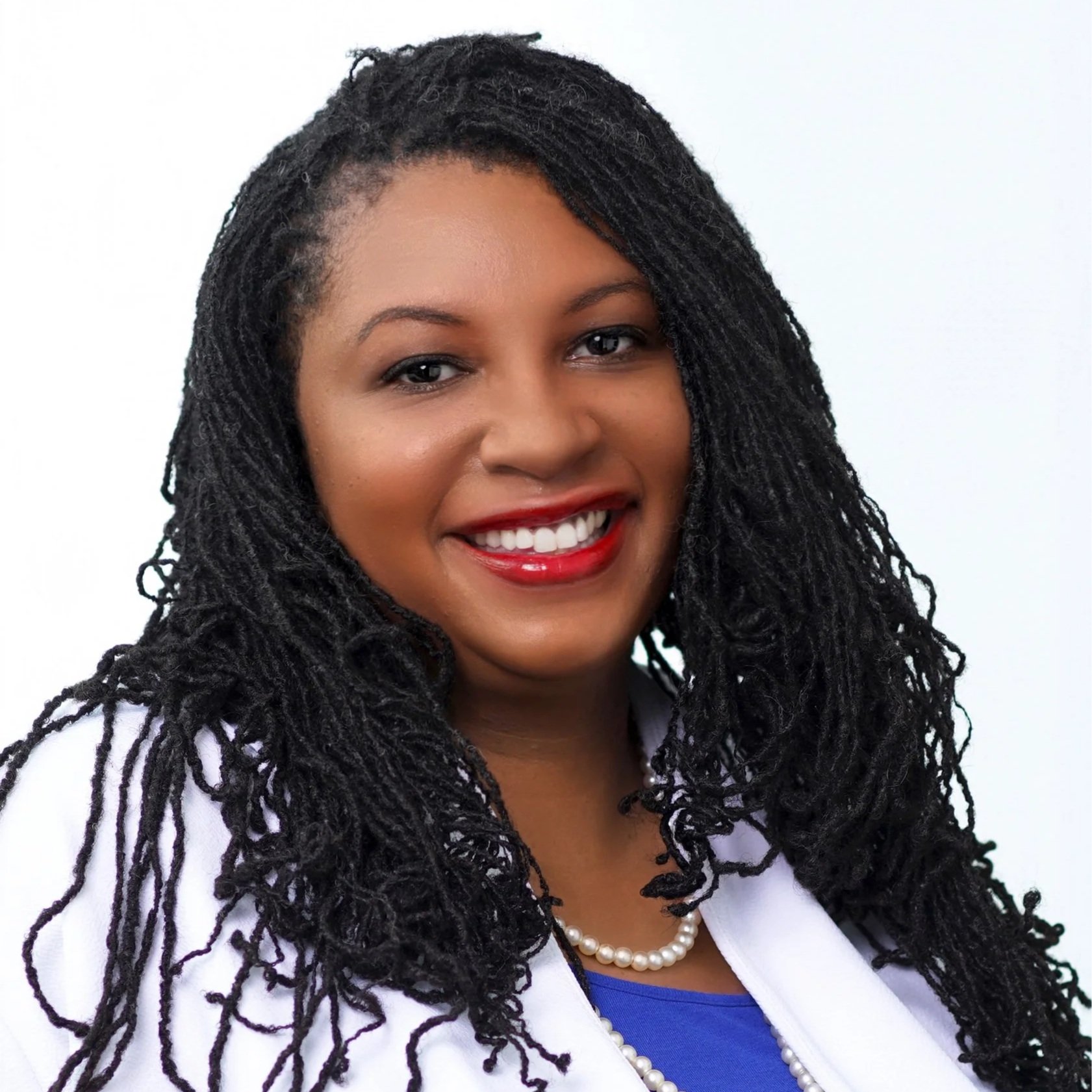 Headshot of a smiling woman with dark hair styled in twists, wearing a white blazer, blue top, pearl necklace, and red lipstick, against a plain white background.