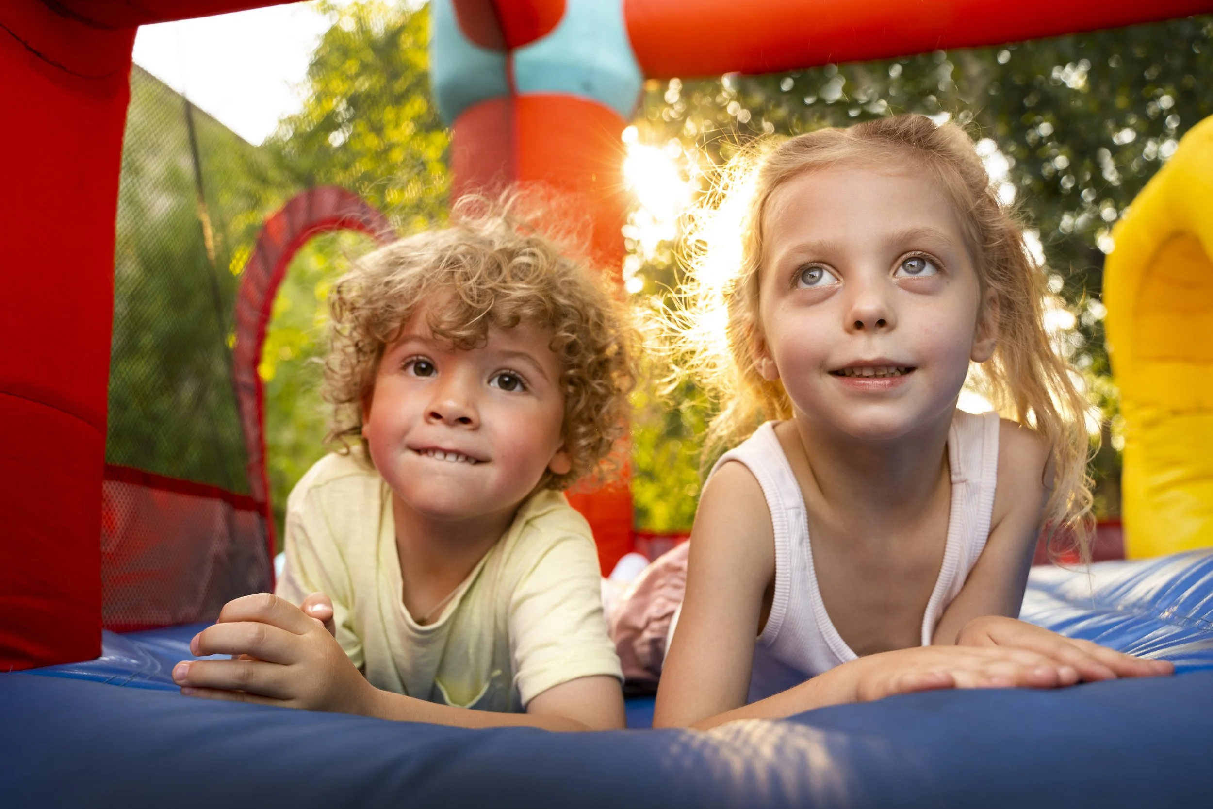 Deux enfants jouent sur un château gonflable dans un parc en plein air, ensoleillé, entourés d'arbres.
