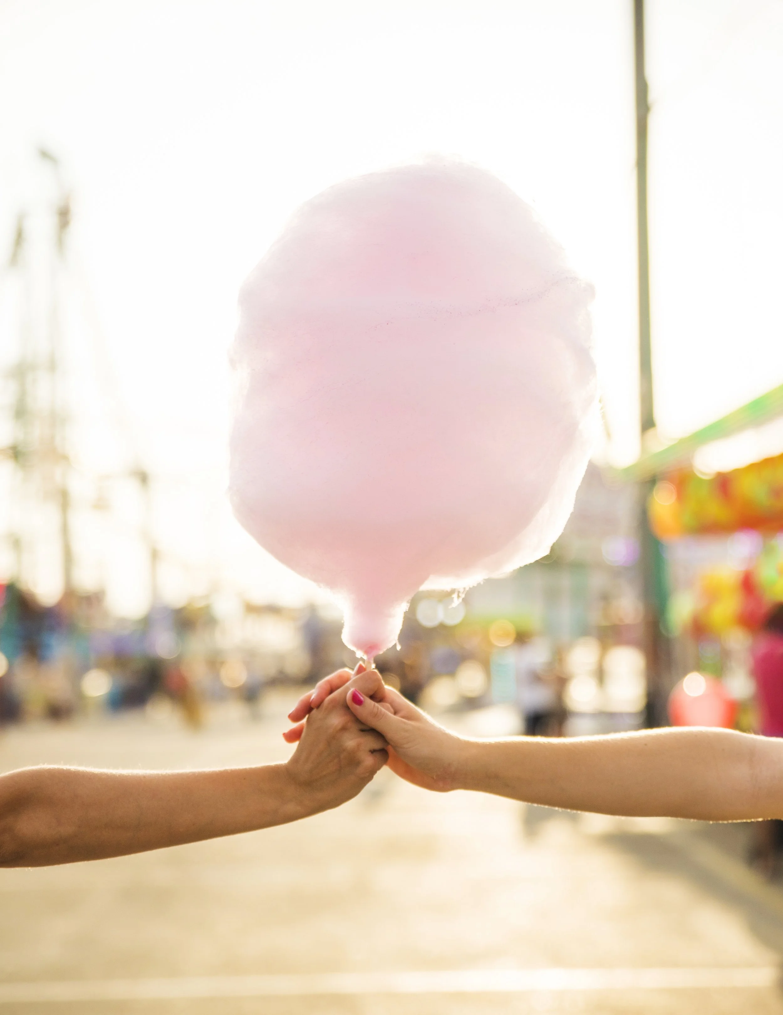 Deux mains tenant une grosse boule de coton de couleur rose pâle, avec un arrière-plan flou d'une fête foraine en plein air, éclairée par la lumière du soleil.