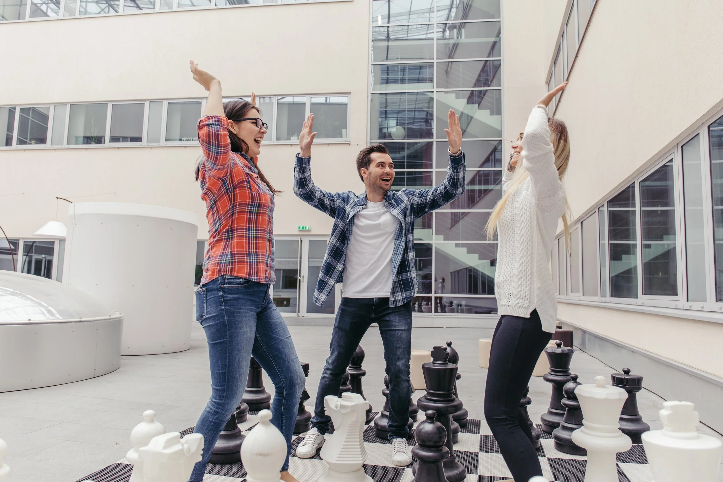 Trois jeunes adultes jouent aux échecs en levant les bras, sur un échiquier géant en plein air à l'extérieur d'un bâtiment moderne.