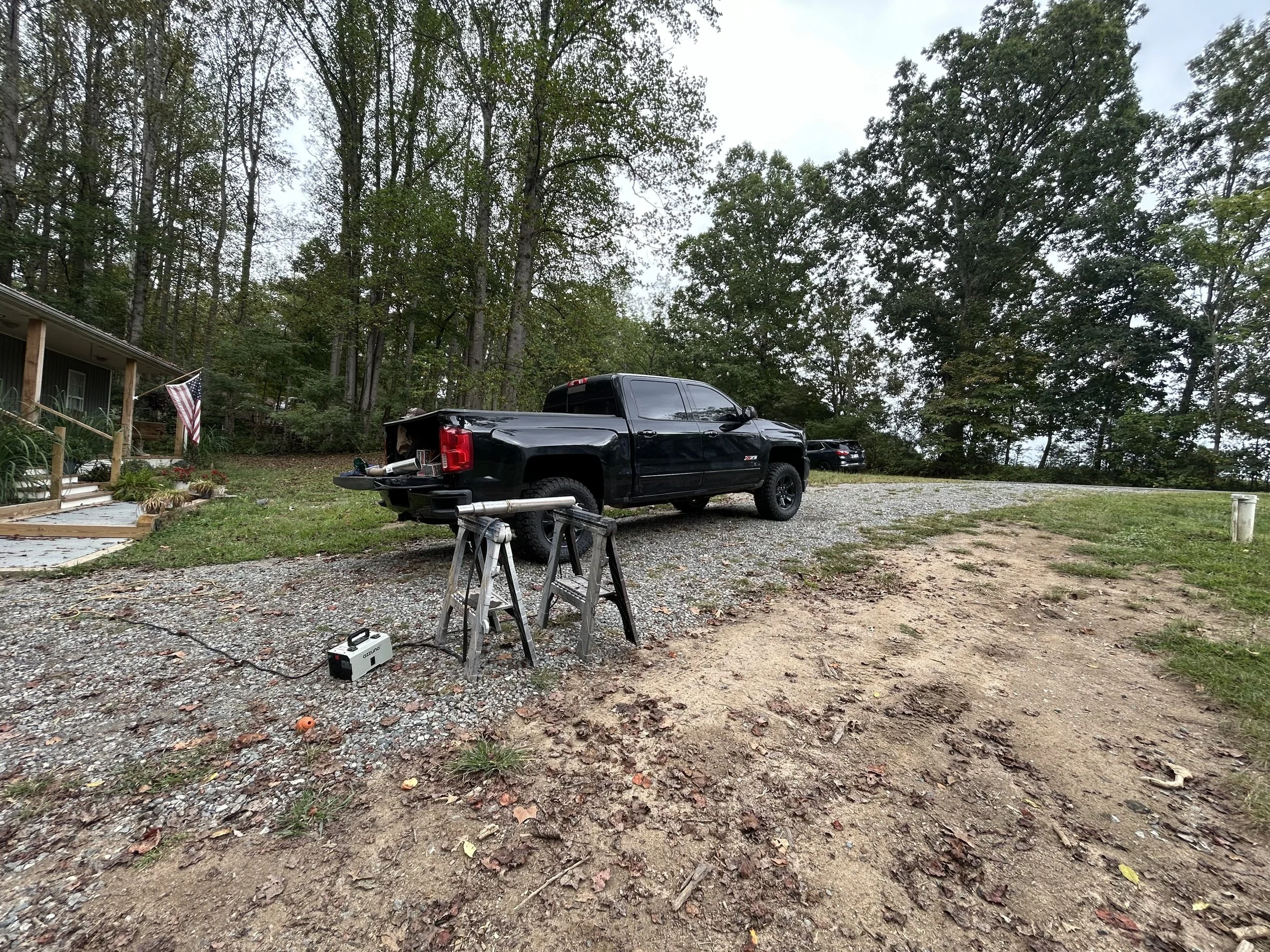 Black pickup truck parked on gravel driveway next to a house, with trees in the background, outdoor work setup with a power tool on a sawhorse, and another vehicle in the distance.