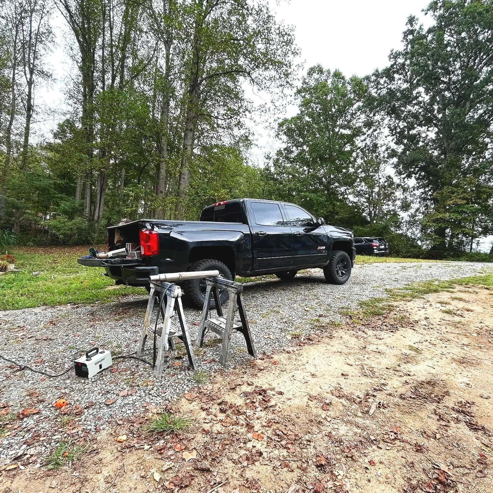 A black pickup truck parked on a gravel driveway next to a wooded area, with a workbench and saw in the foreground.