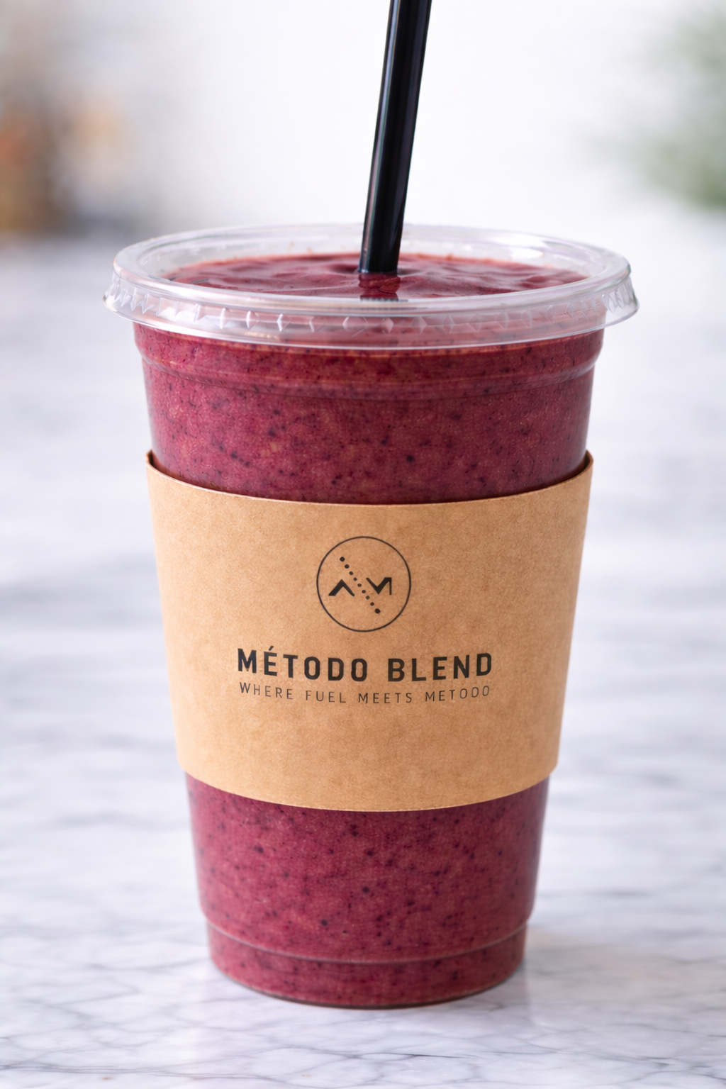 A close-up of a blended berry drink in a clear plastic cup with a black straw, wrapped with a brown paper sleeve with the logo 'Método Blend' and the slogan 'Where fuel meets metodo,' on a marble surface.