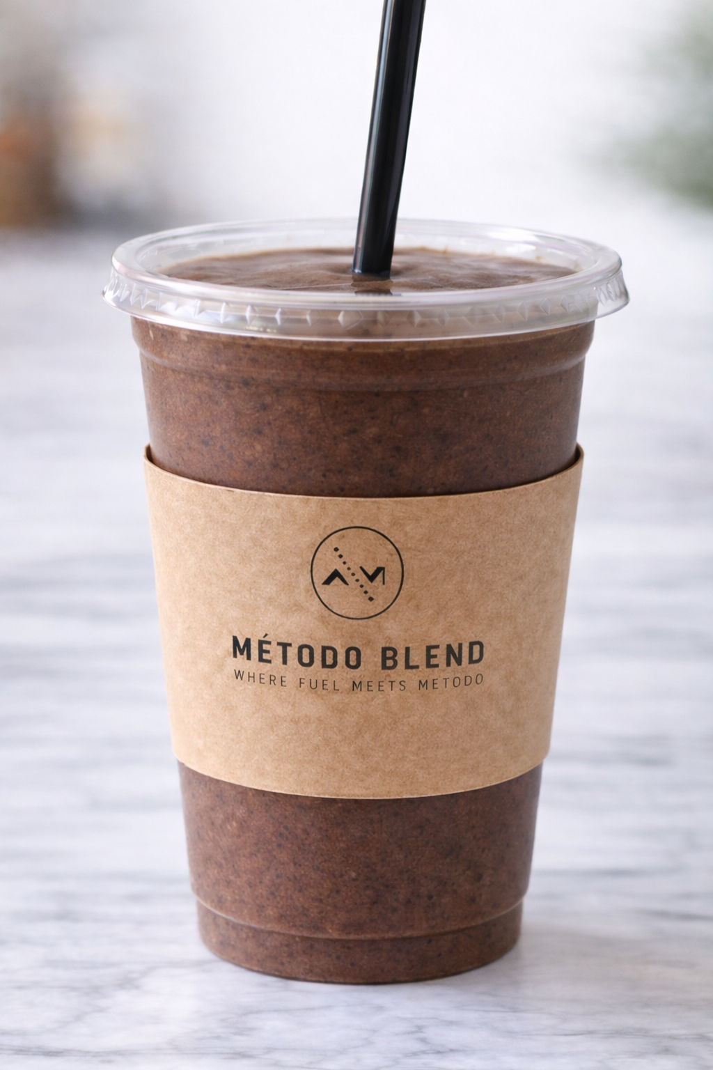 A cold coffee drink in a plastic cup with a black straw, wrapped with a brown paper sleeve labeled 'Método Blend' and 'Where fuel meets metodo,' placed on a light-colored surface.